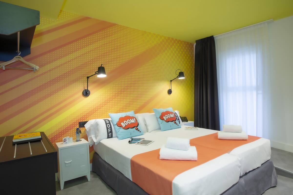 A hotel room with a bed, two bedside tables, a desk, a chair, a window with curtains, and a wall with a geometric pattern.
