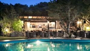 A poolside area with a bar and seating, surrounded by trees and a rocky landscape. - Hotel Rocce Sarde (Olbia)