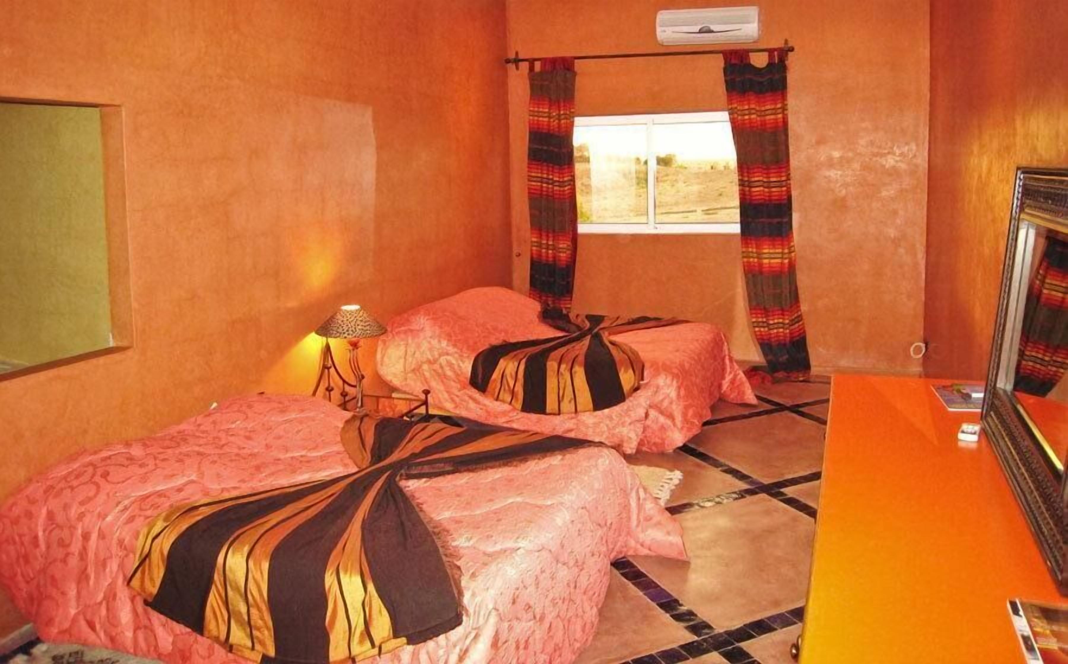 Una habitación con dos camas, una mesita, una lámpara y un ventanal con cortinas.