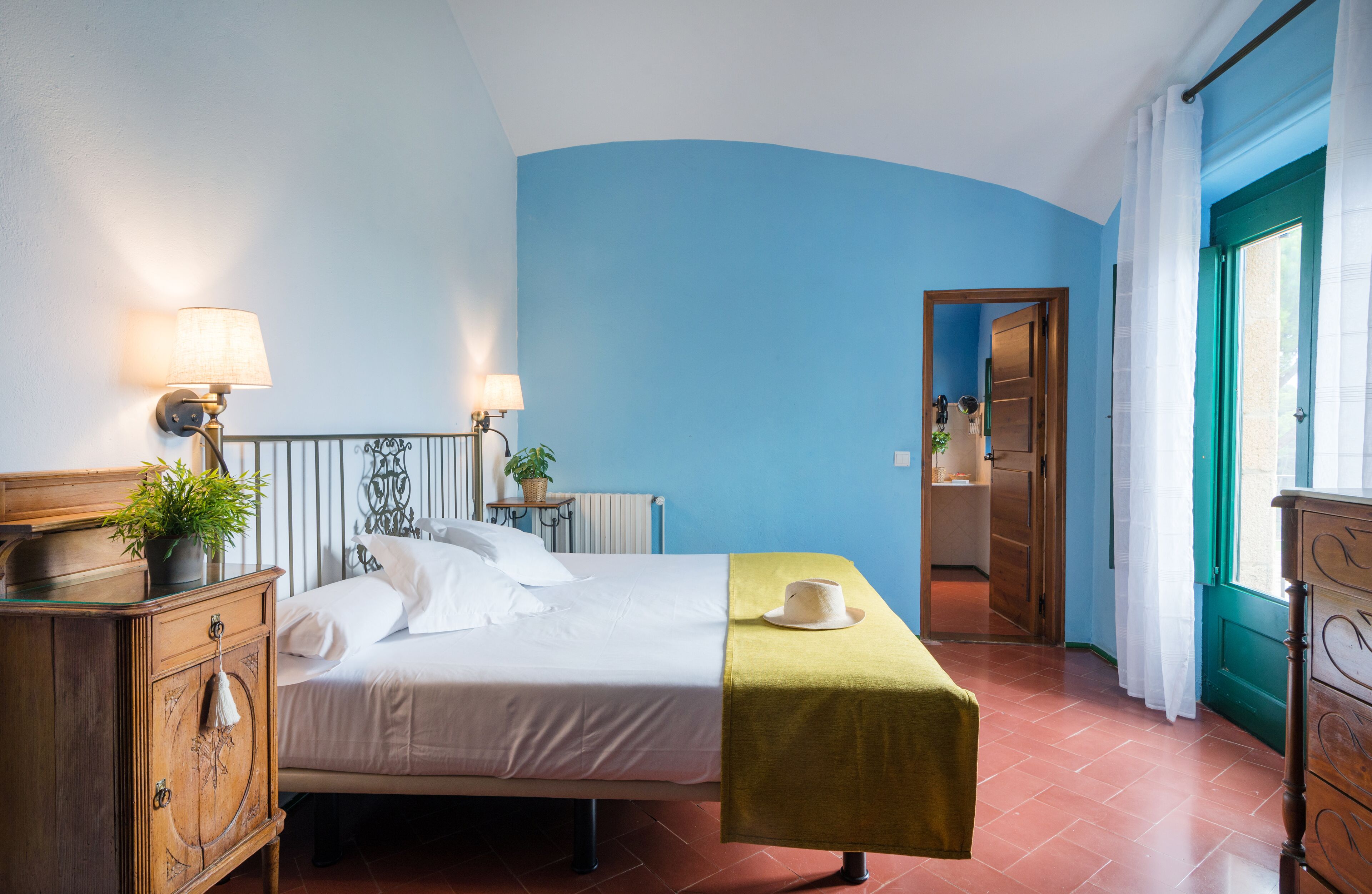 Un dormitorio con cama, lámparas de noche, cómoda, ventana con cortinas y puerta que da a otra habitación.