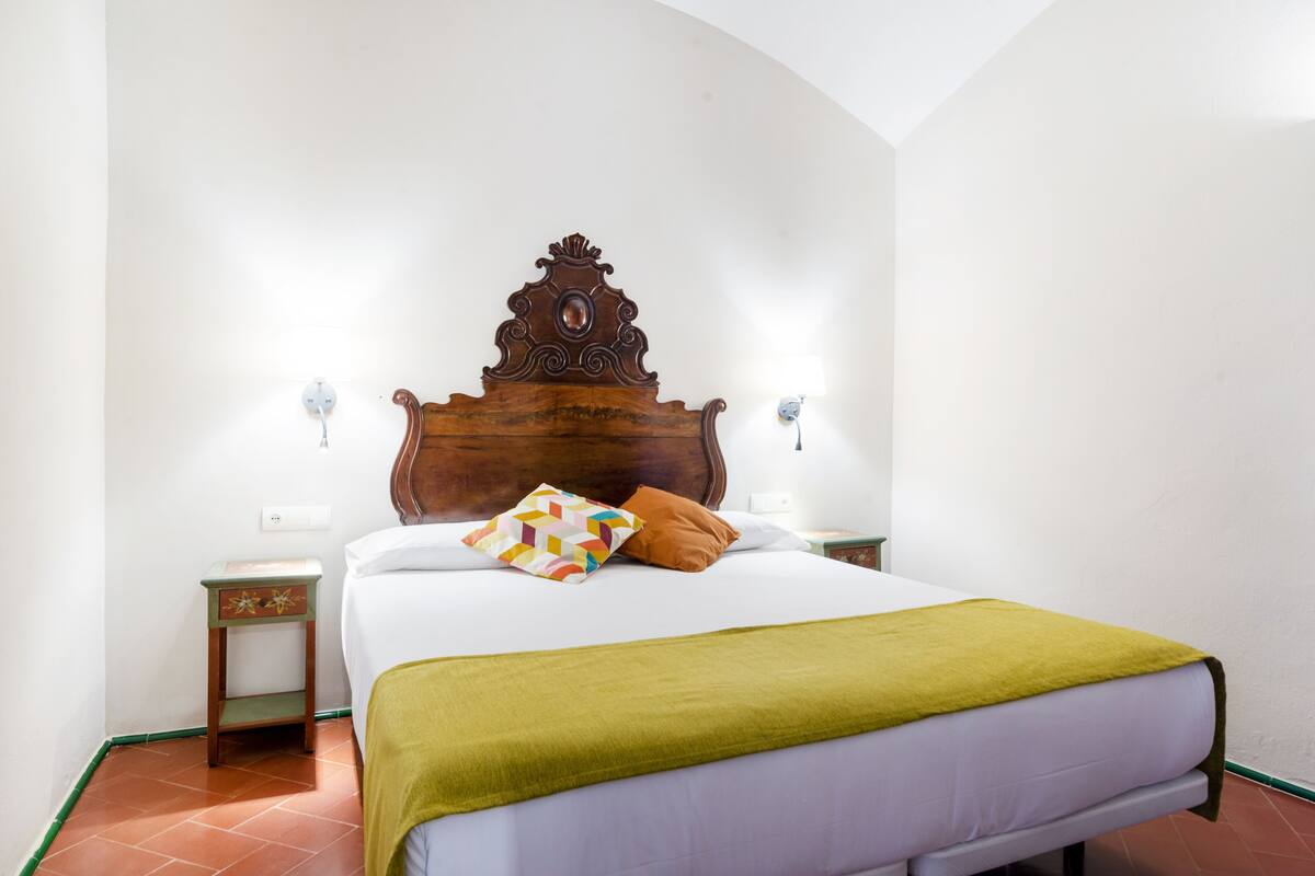 Un lit bien fait, avec une tête de lit en bois, un plaid vert et deux coussins décoratifs. Il y a deux tables de chevet avec des lampes, et la pièce a des murs blancs et un sol carrelé.