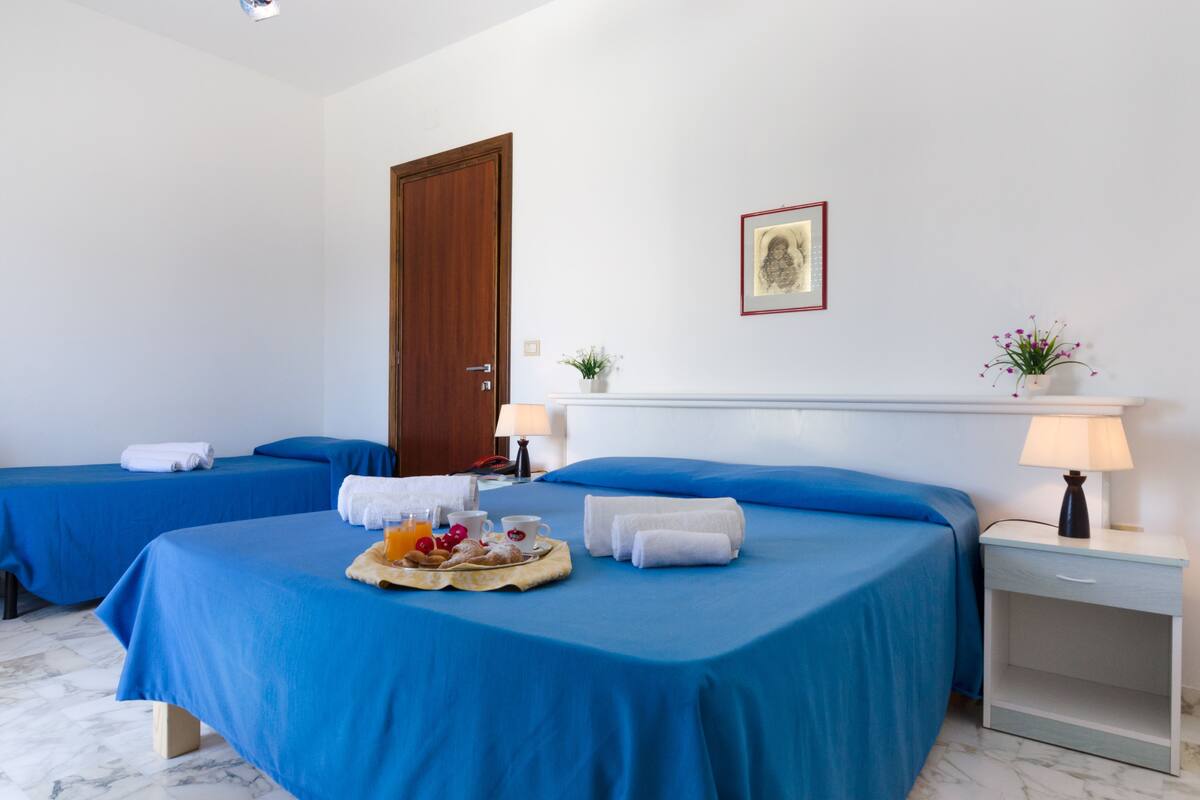 A hotel room with two beds, a wooden door, a framed picture, a nightstand with a lamp, and a tray with breakfast items.