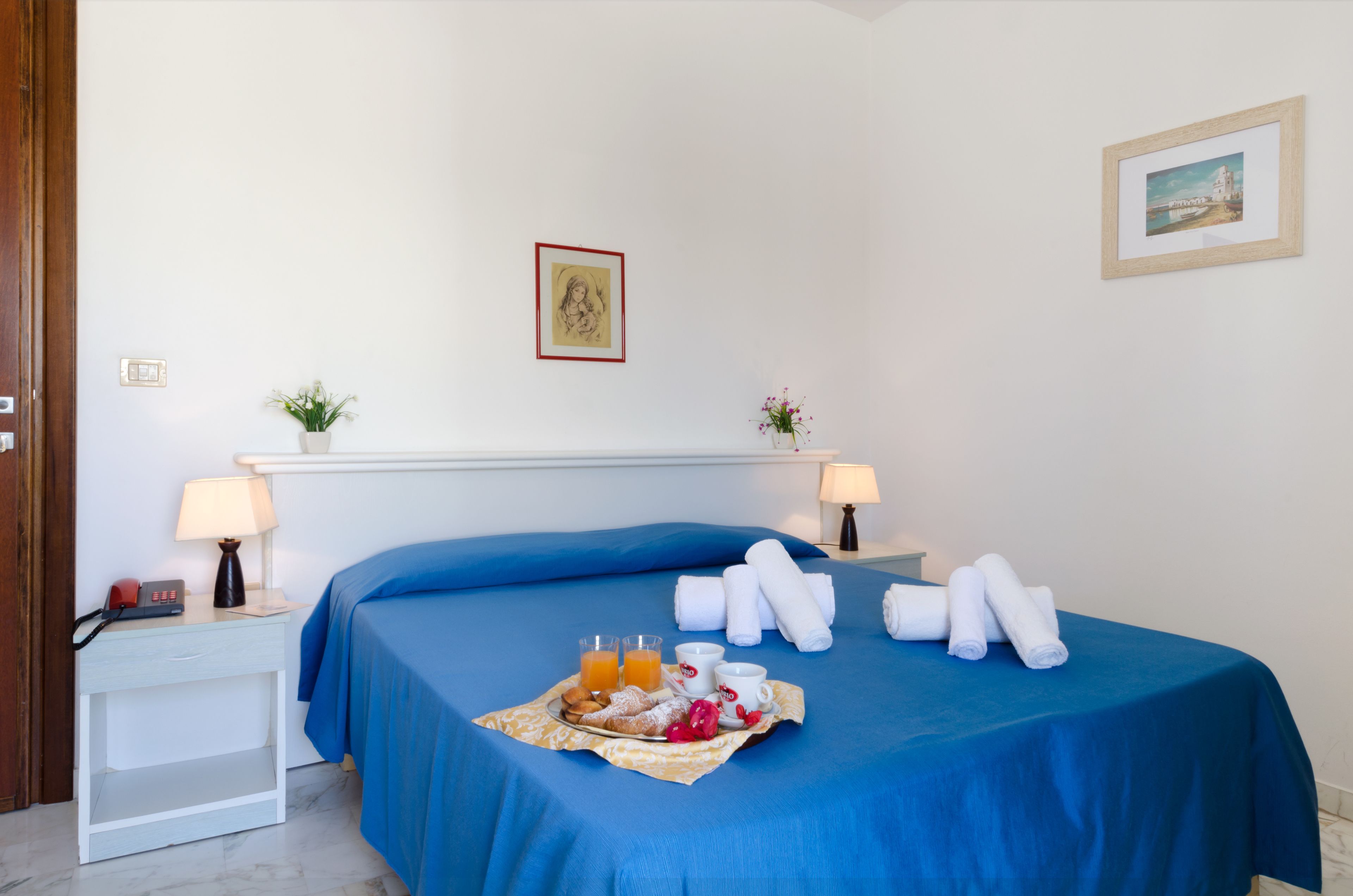 A hotel room with a blue bedspread, a breakfast tray with pastries, juice, and coffee, and a small table with a lamp and a telephone.