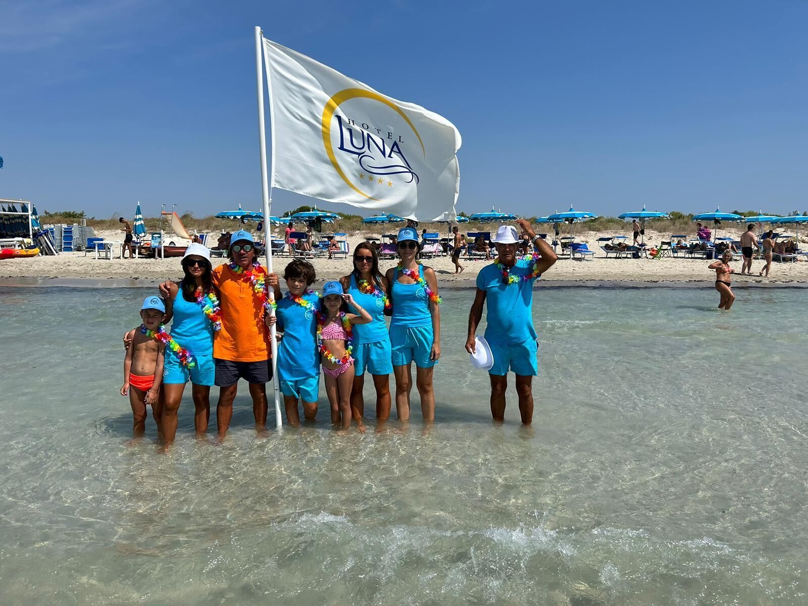 Un groupe de personnes se tient dans une eau peu profonde sur une plage, et on peut voir un drapeau avec l’inscription « HÔTEL LUNA ».