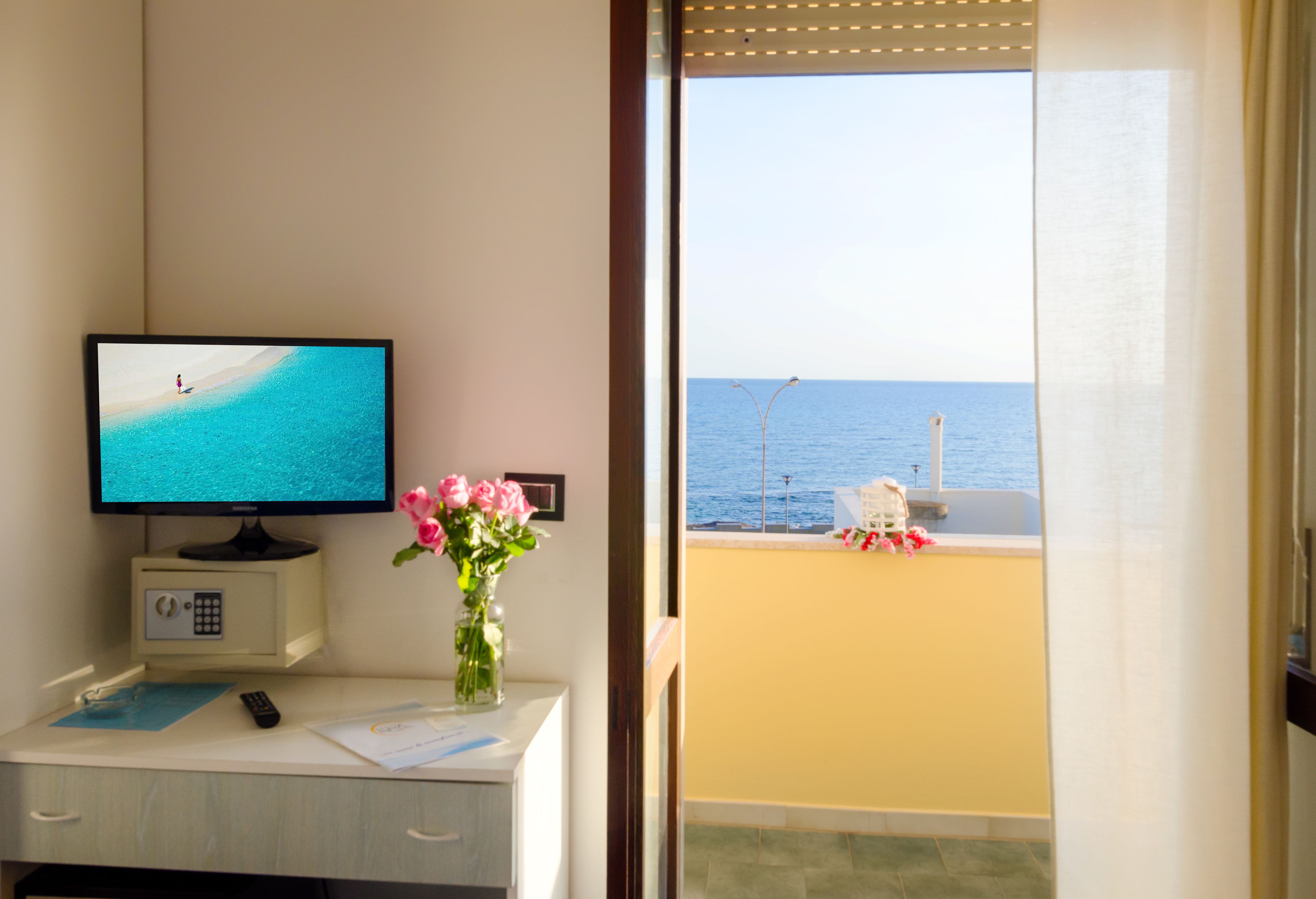 A room with a TV on a desk, a vase of flowers, and a view of the sea through a balcony door.