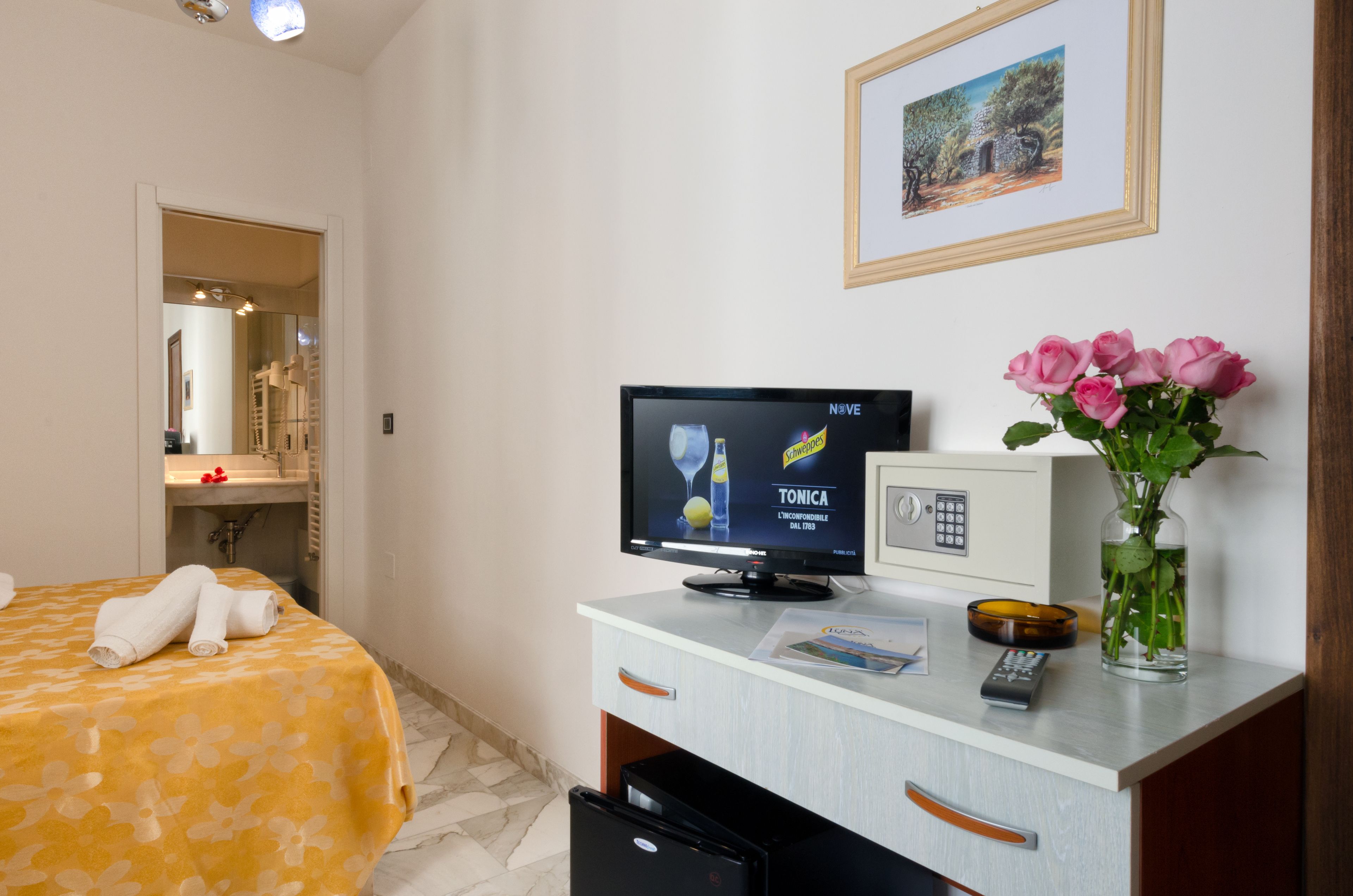 Une chambre d’hôtel avec un lit, une télévision, une télécommande, un vase de fleurs et un tableau encadré au mur.