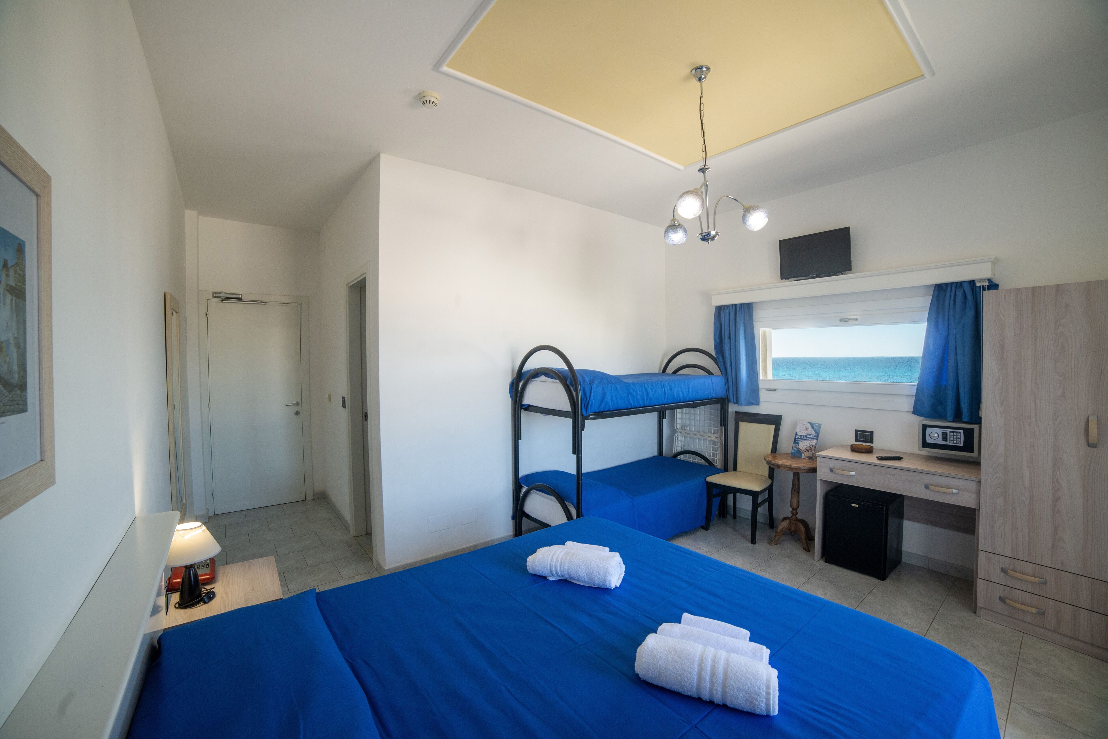 A room with bunk beds, a desk, a chair, a small fridge, a lamp, and a television.