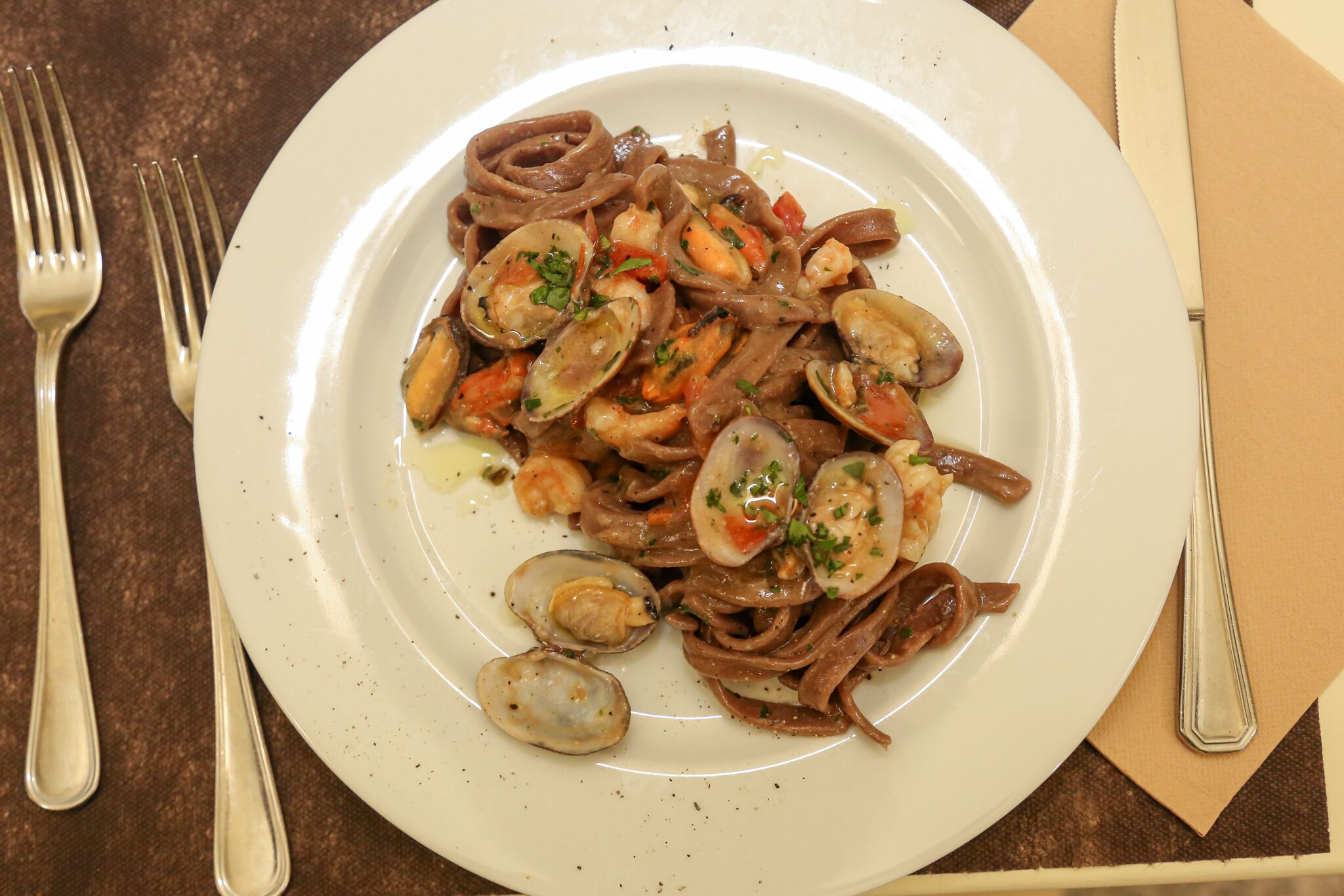 En tallerken med skaldyrs pasta med muslinger og rejer, ledsaget af en gaffel og kniv på et bord.