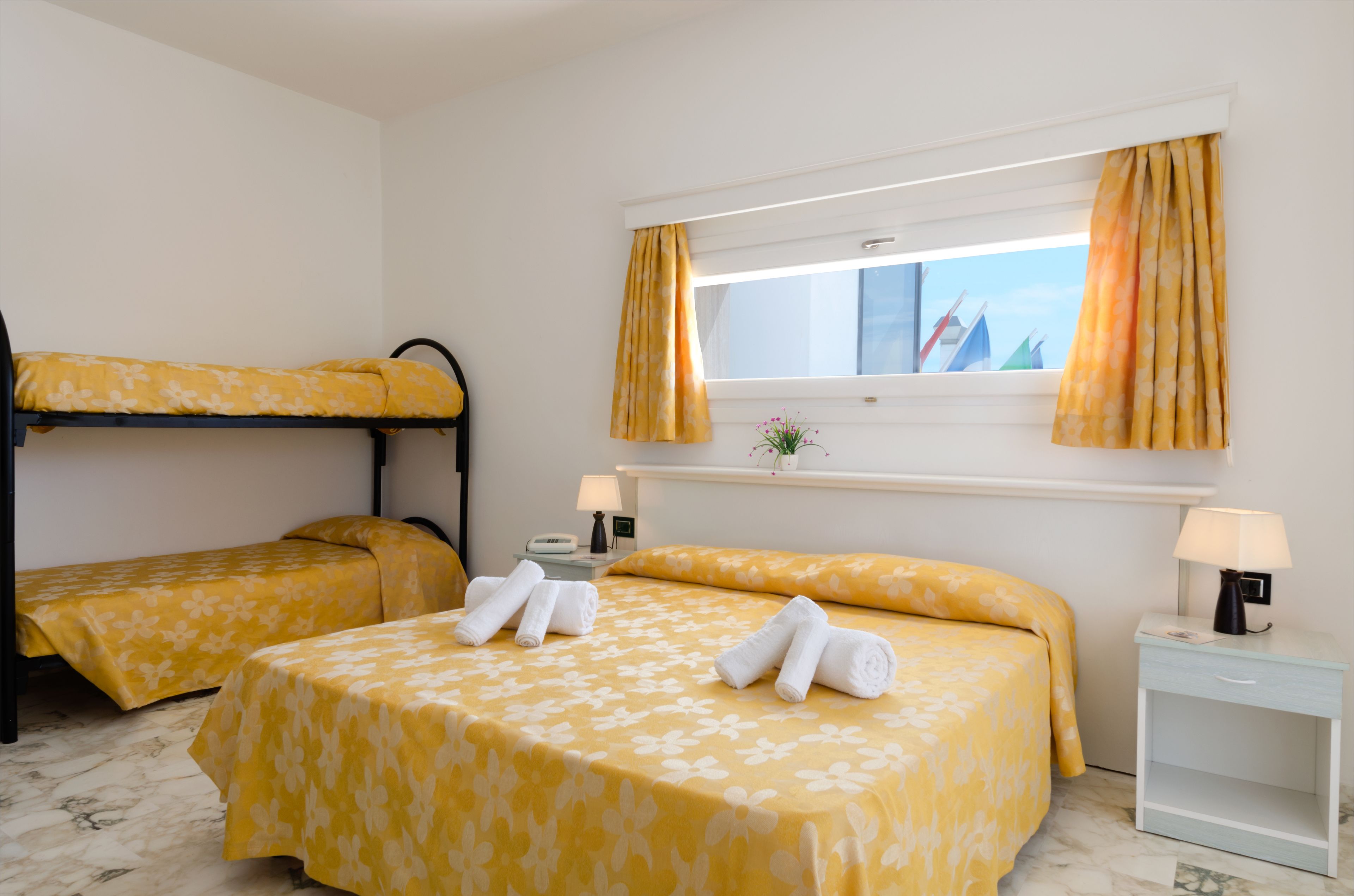 A hotel room with two beds, a window with curtains, a nightstand with a lamp, and a small table with a vase of flowers.