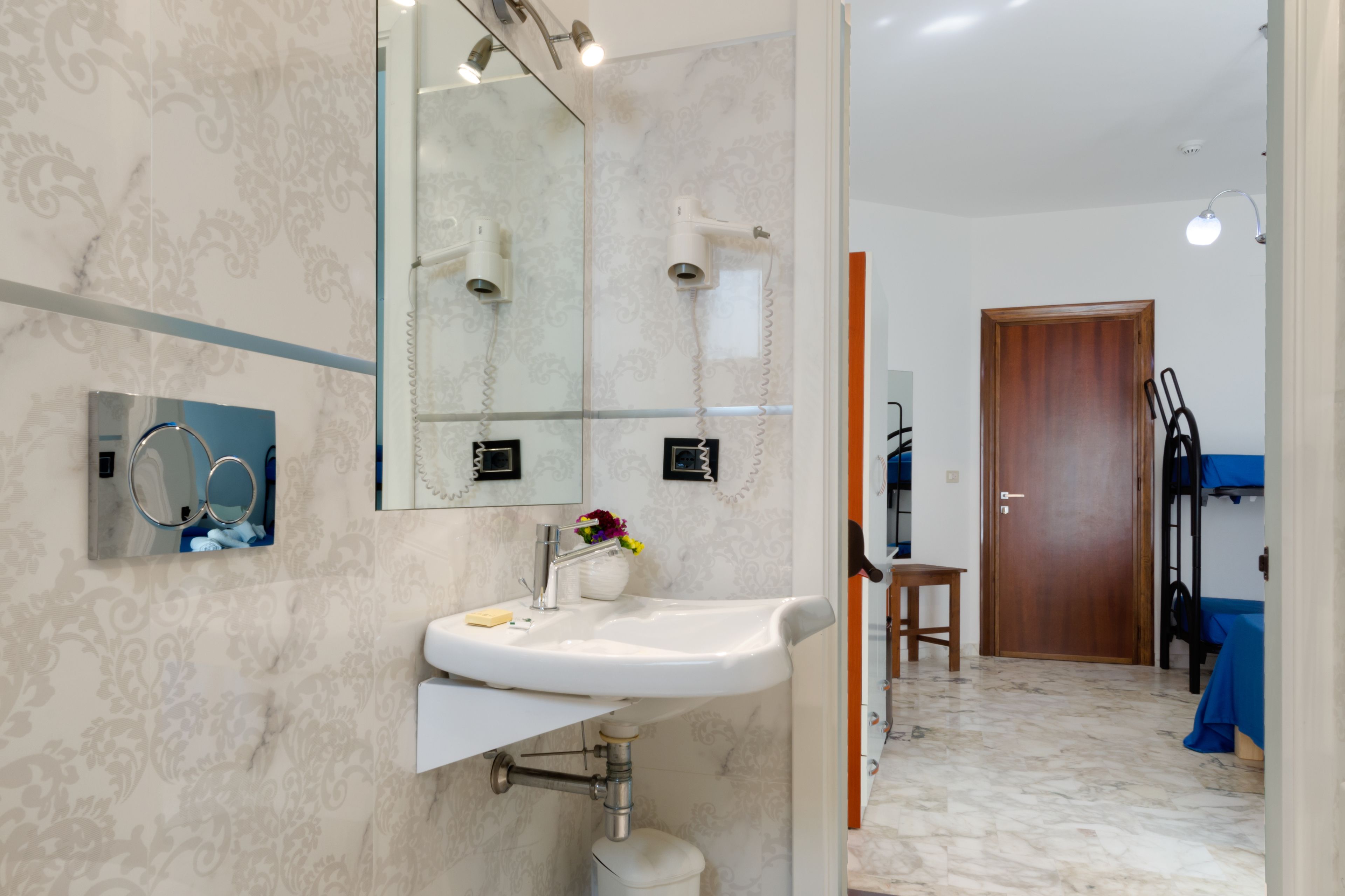 A modern bathroom with a large mirror, a sink, and a shower. There is a wooden door and a small table in the adjacent room.