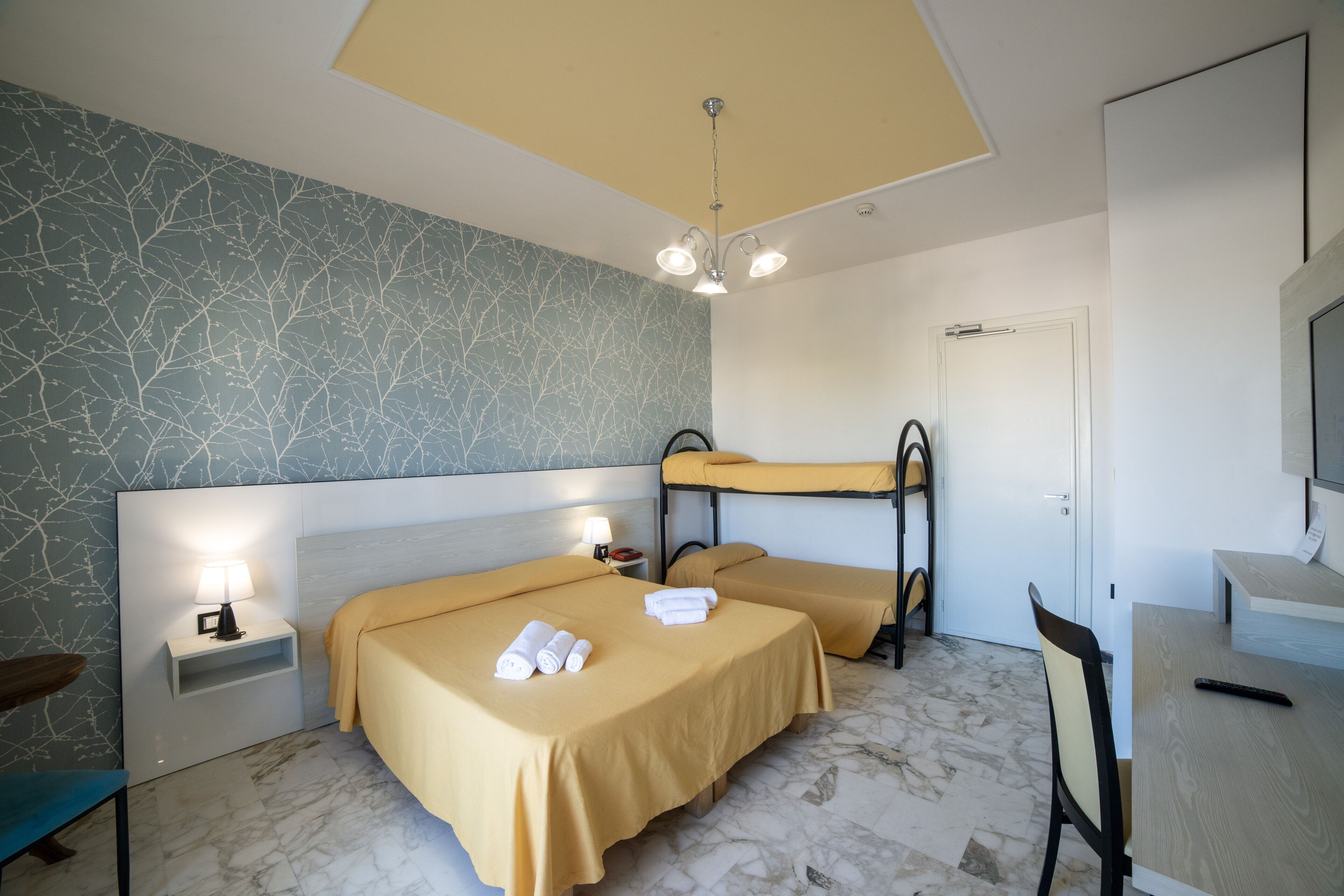 A hotel room with two beds, a desk, a chair, a nightstand with a lamp, and a ceiling light fixture.
