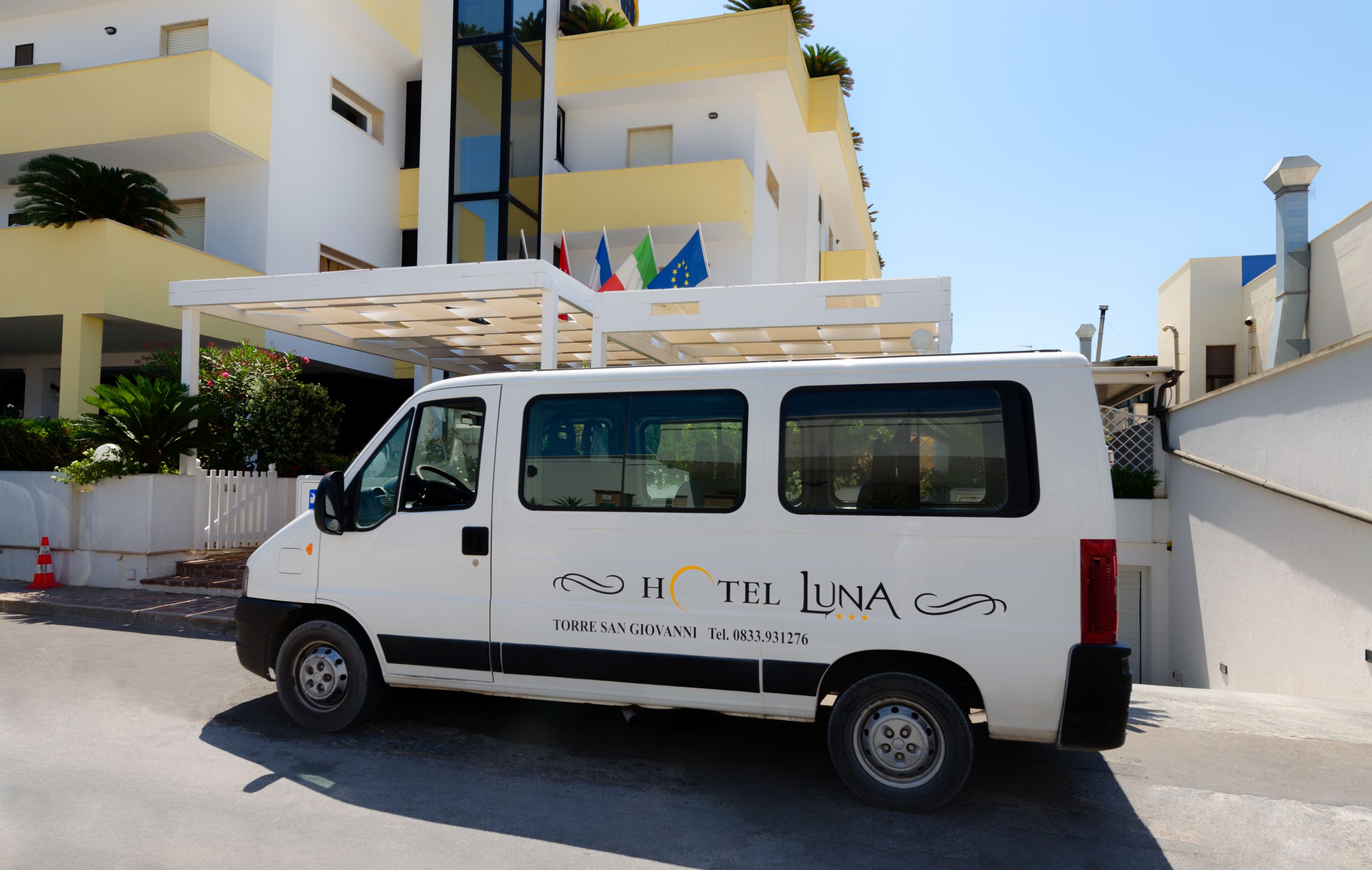 A white van parked in front of a building with balconies and flags.