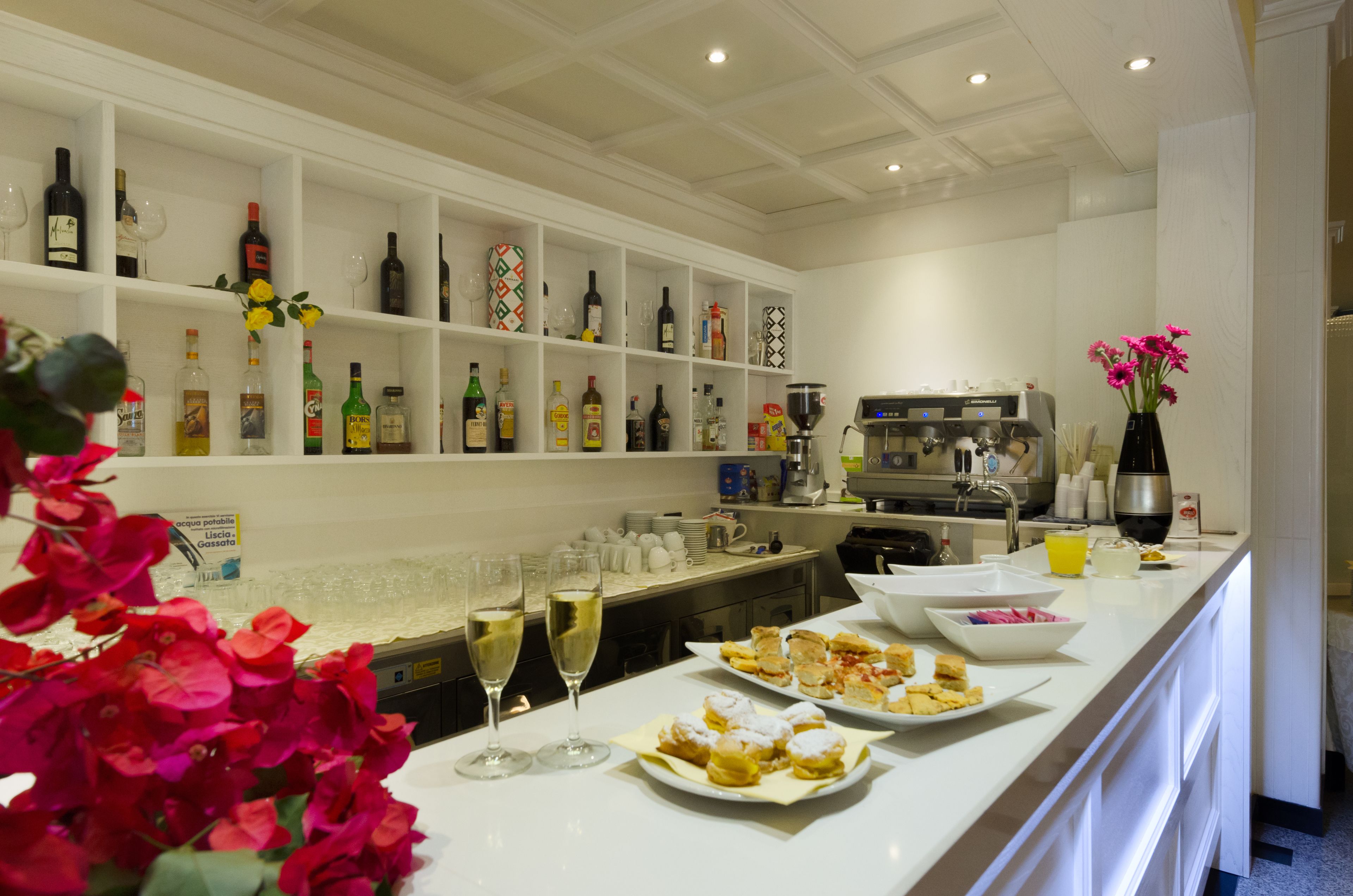 Una cocina de hotel con estantes blancos repletos de botellas y copas, una máquina de café y un mostrador con platos de comida y vasos de vino.