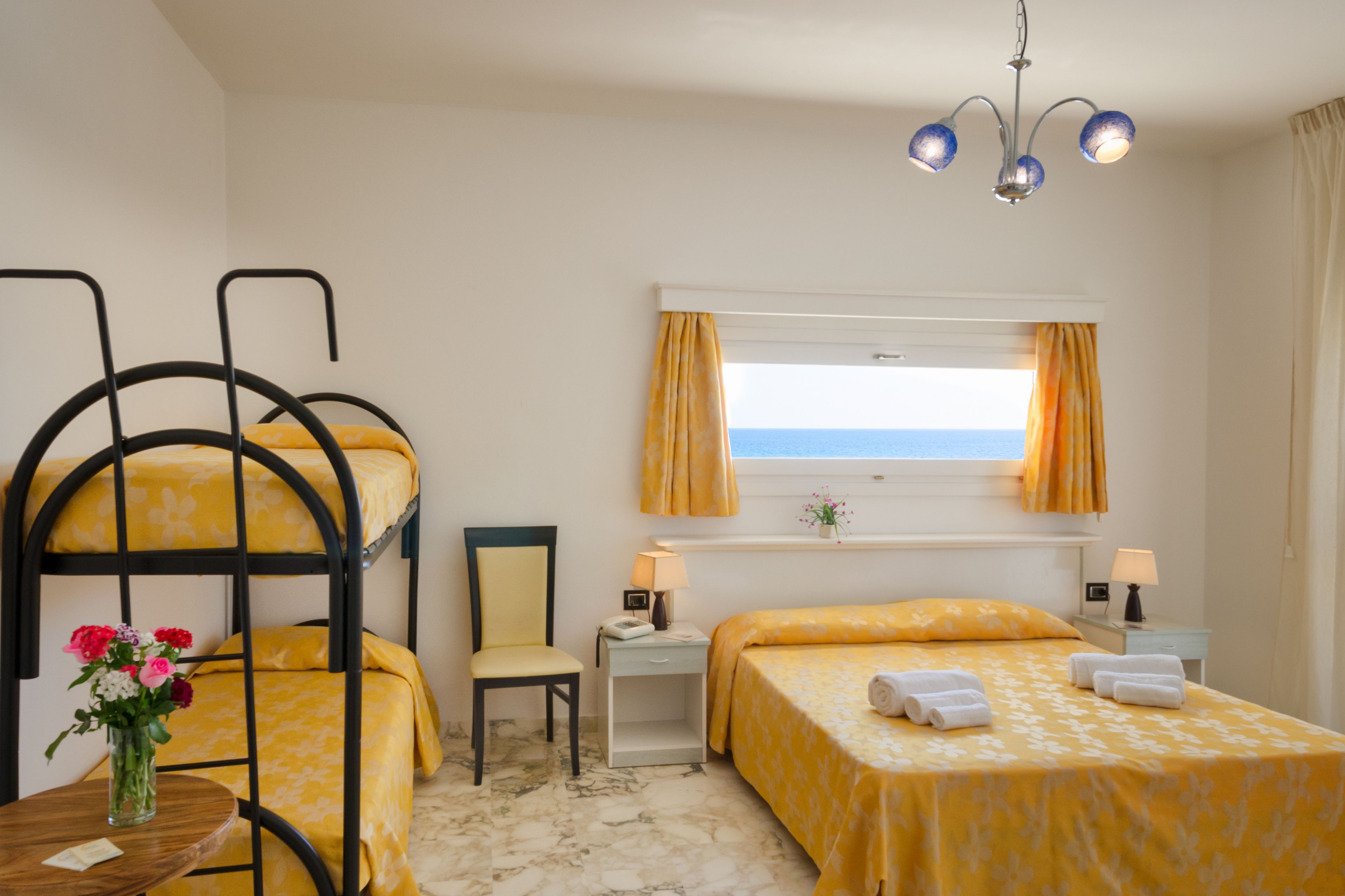 A hotel room with bunk beds, a single bed, a chair, a small table with a vase of flowers, a window with curtains, and a ceiling light fixture.