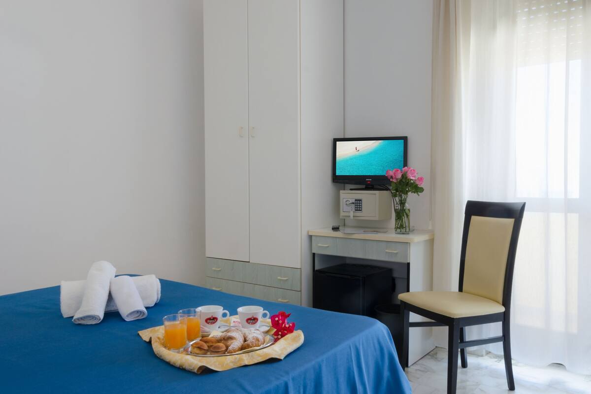 A hotel room with a bed, a desk with a computer, a chair, and a breakfast tray with pastries and juice.
