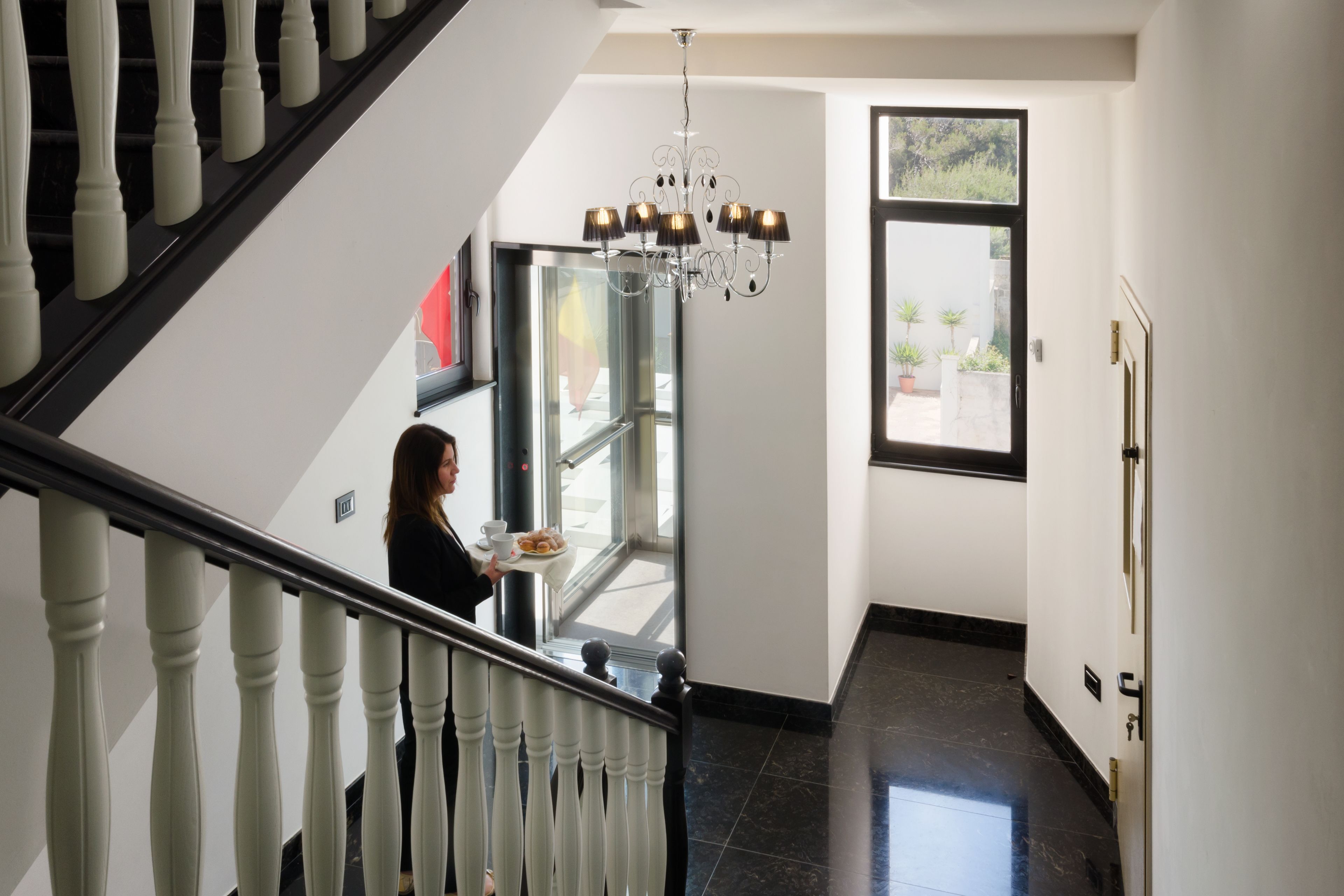 A staircase with white balusters, a chandelier, and a person holding a tray with food.