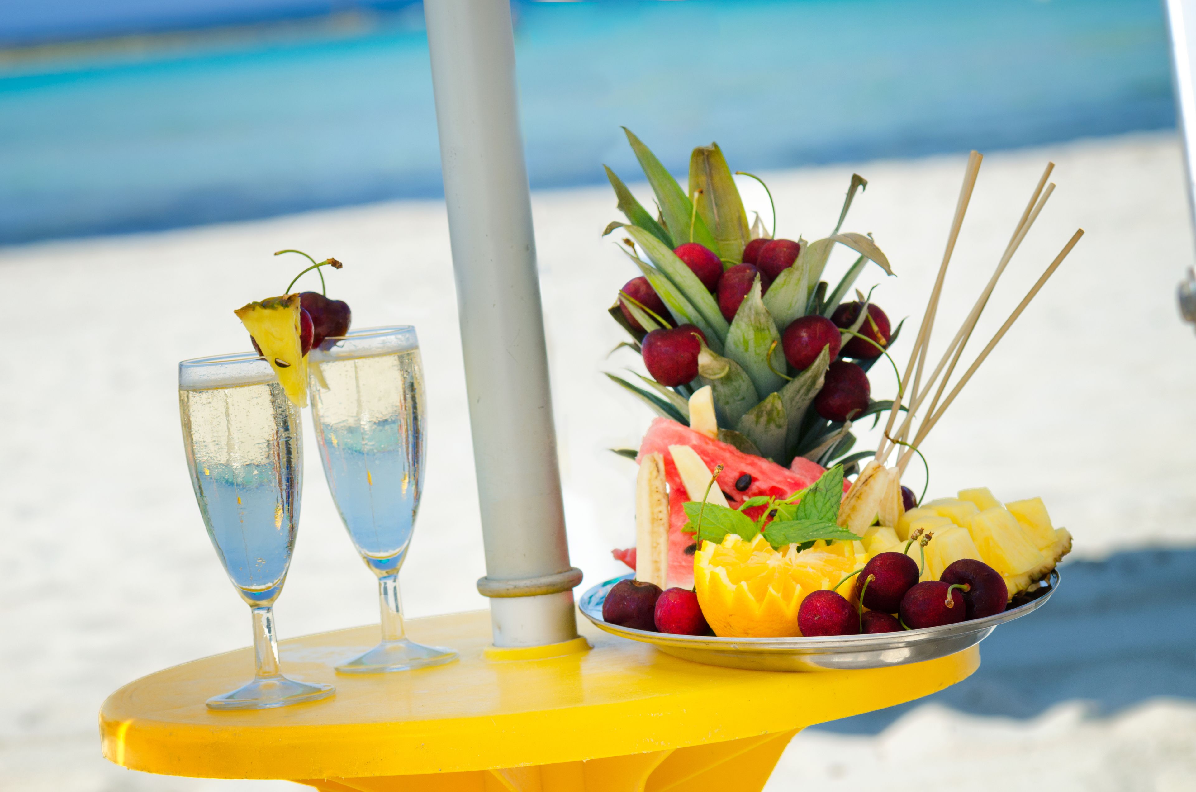 Une table avec deux verres contenant une boisson claire, une assiette de fruits comprenant de la pastèque, de l’ananas et des cerises, ainsi qu’une composition décorative de baies rouges et de feuilles vertes.