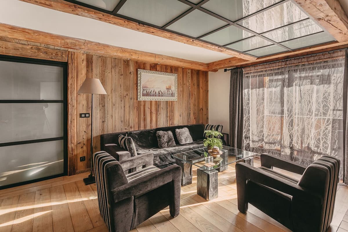 Ein modernes Wohnzimmer mit Holzvertäfelung, einem schwarzen Sofa, einem Glastisch und einem großen Fenster mit Vorhängen.
