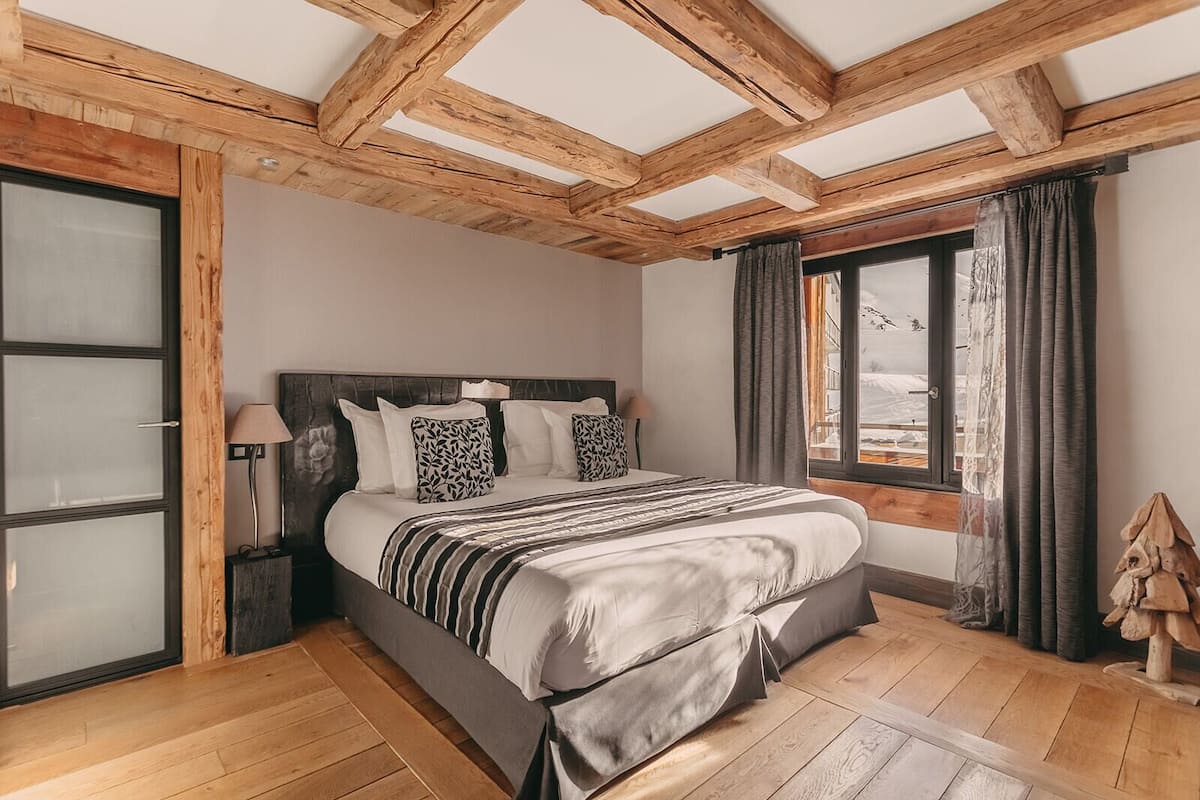 Ein Schlafzimmer mit Holzbalkendecke, einem großen Bett mit gemusterten Kissen, einem Nachttisch mit Lampe und einem Fenster mit Vorhängen.