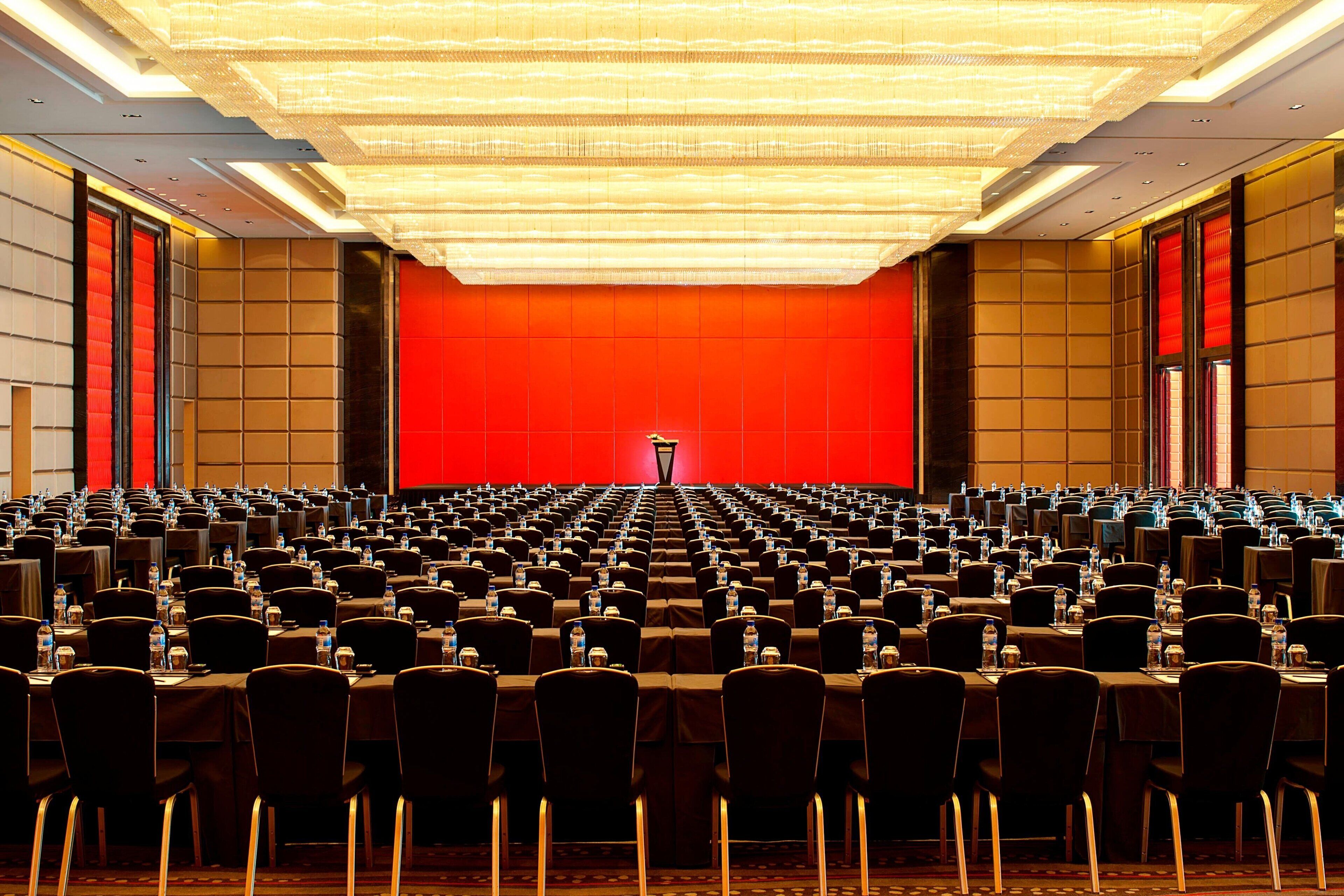 Une salle de conférence avec des rangées de tables et de chaises, un pupitre à l’avant et un mur rouge.