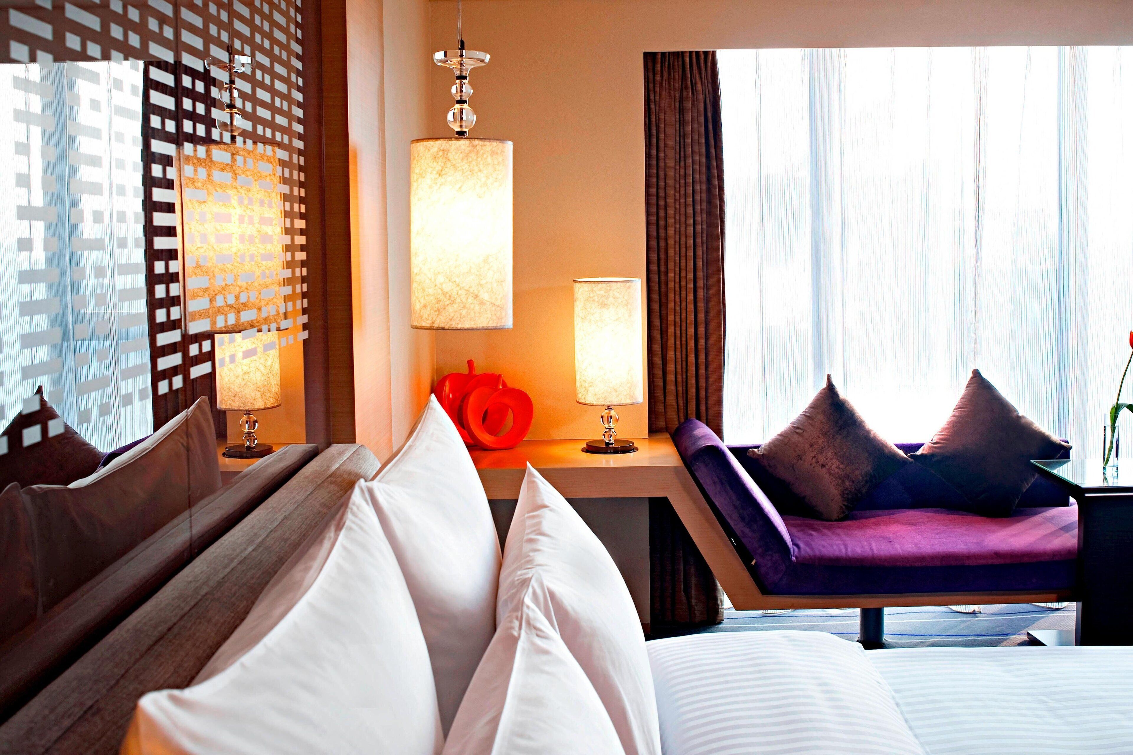 Ein Hotelzimmer mit Bett, Sofa, Schreibtisch mit Lampe und Blick auf die Stadt durch das Fenster.
