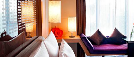 A hotel room with a bed, a sofa, a desk with a lamp, and a view of the city through the window.