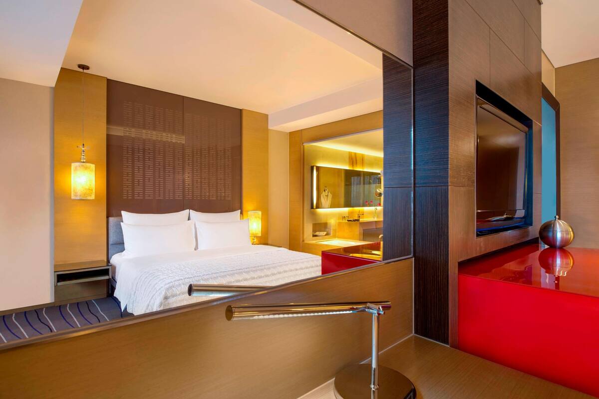 A modern hotel room with a large bed, a bedside table, a mirror, and a red accent wall.