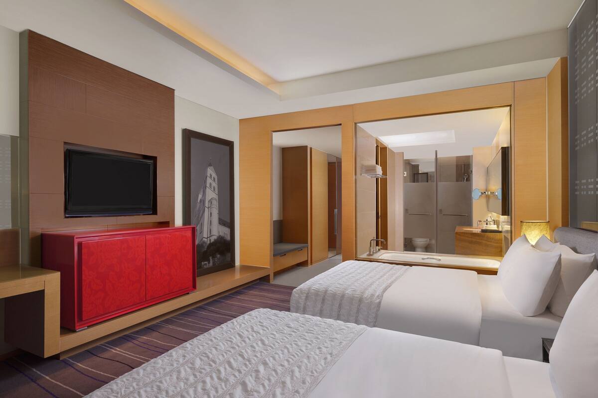 A hotel room with a TV, a red cabinet, two beds, a bathroom, and a mirror.