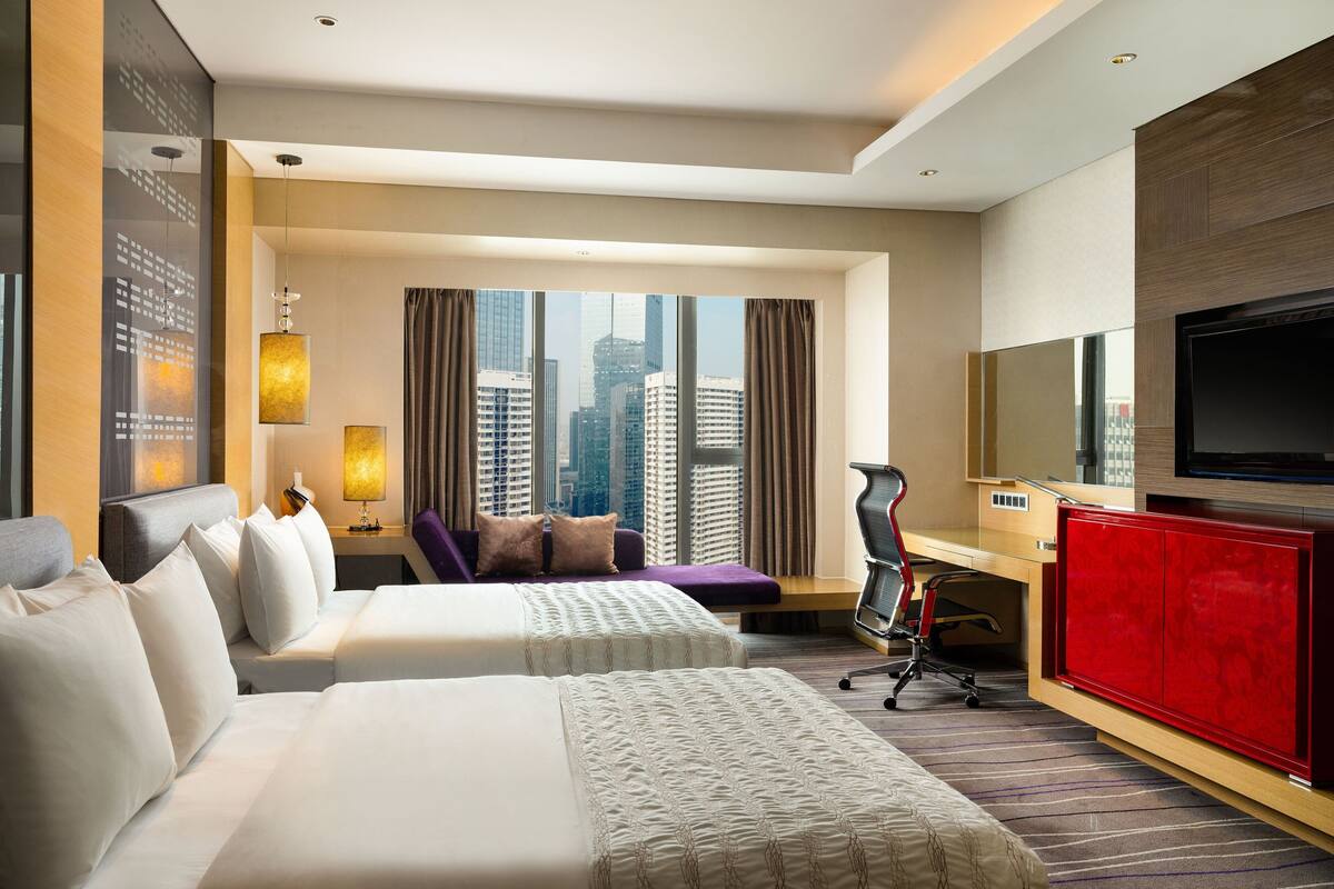 A hotel room with two beds, a desk, a TV, and a large window with a city view.