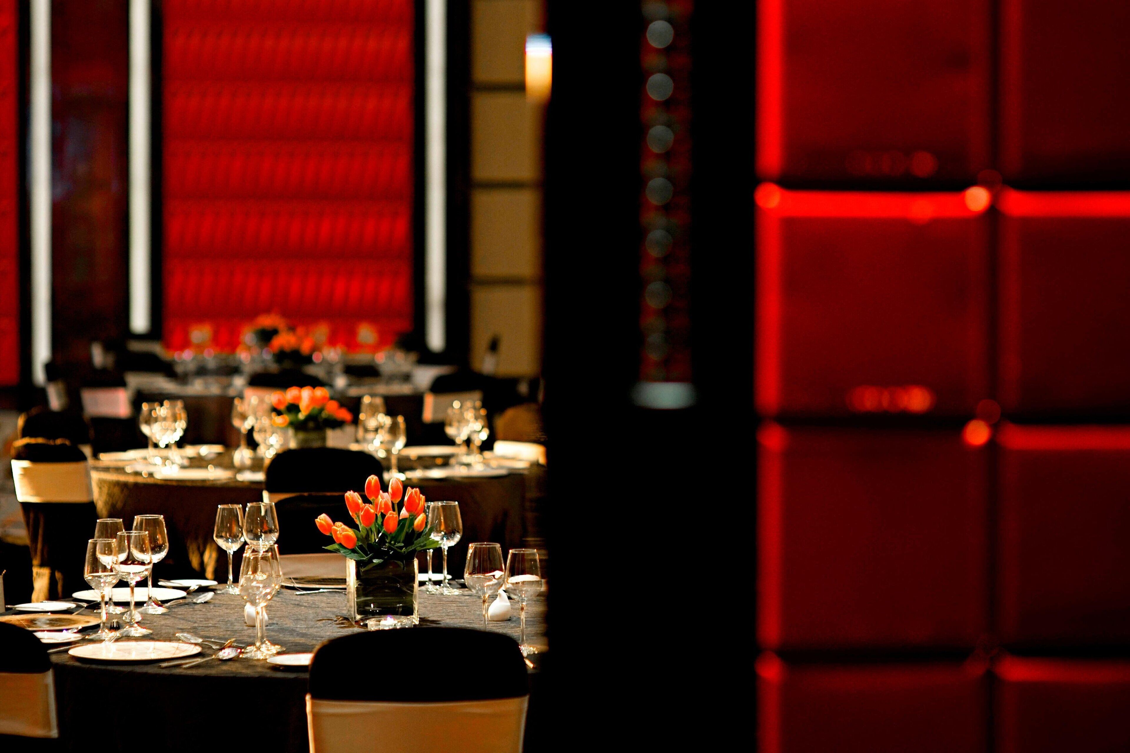 A dining area with tables set for a meal, featuring a vase of orange tulips and wine glasses.