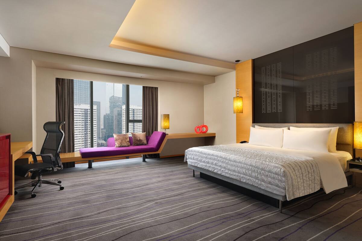 A hotel room with a large bed, a desk, a purple sofa, and a city view.