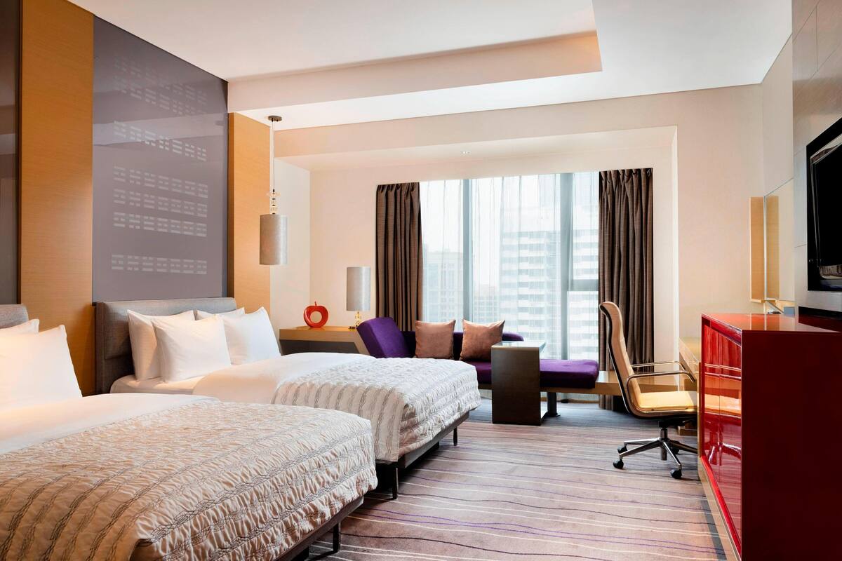 A hotel room with two beds, a desk, a chair, a TV, and a large window with curtains.