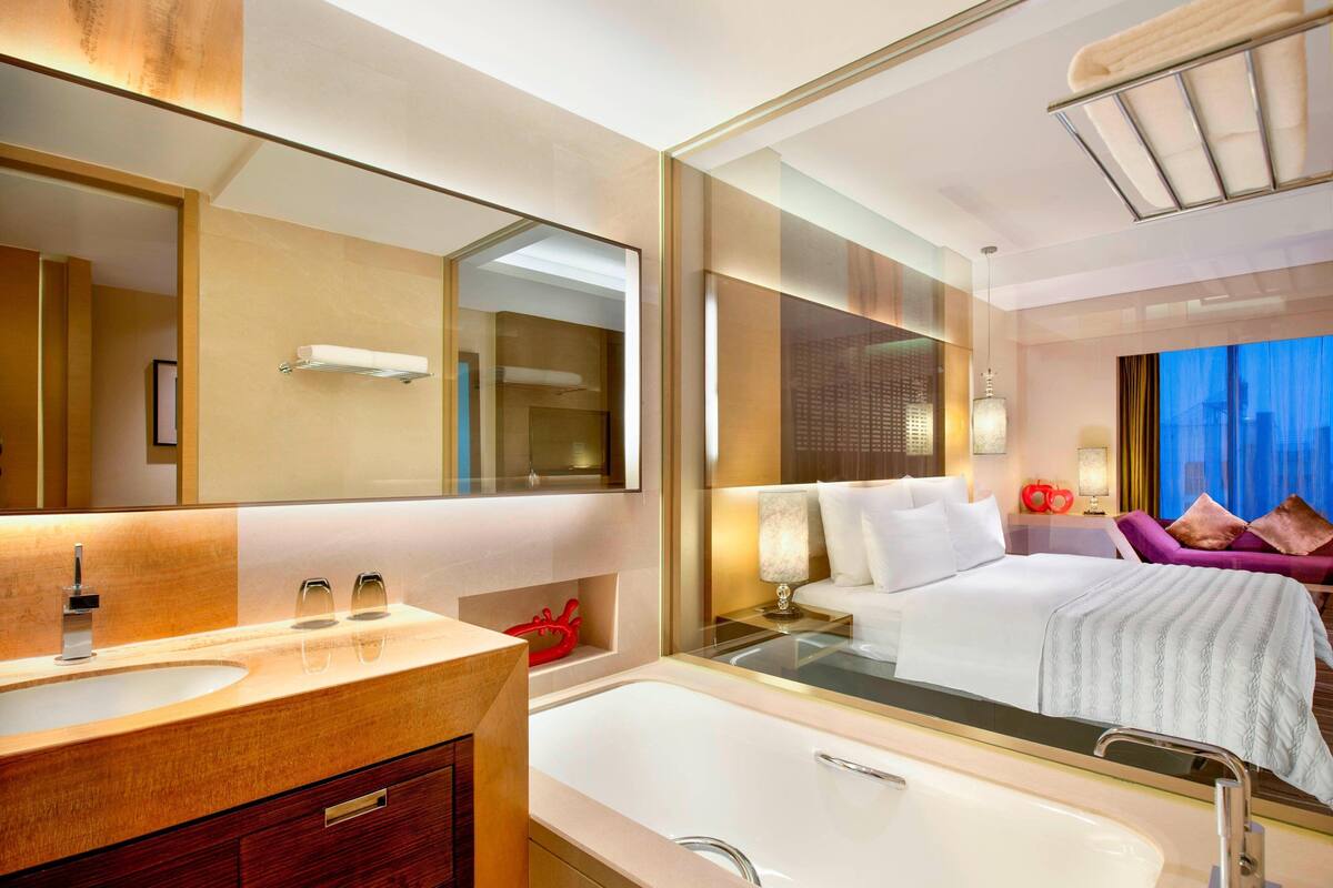 A modern hotel room with a large mirror, a bathtub, a vanity with a sink, a bed with white linens, and a view of the city at night.