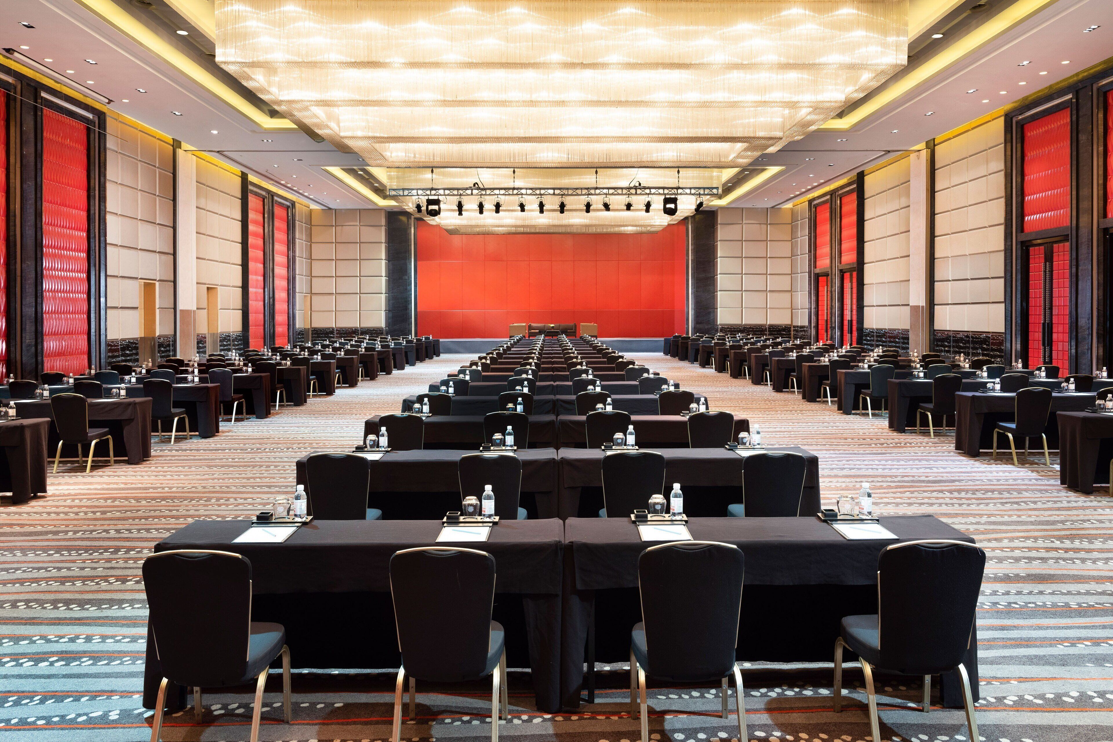 A large conference room with rows of tables and chairs, a stage, and a red backdrop.
