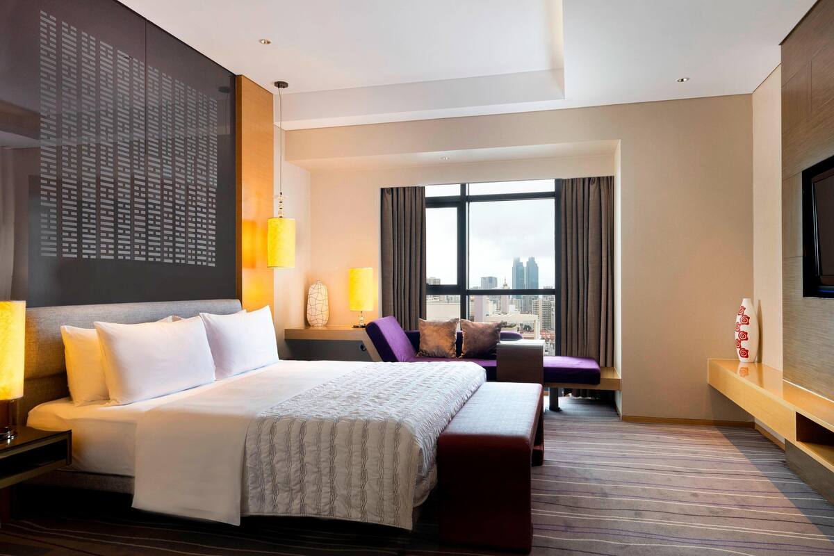 A modern hotel room with a large bed, a seating area, a TV, and a city view.