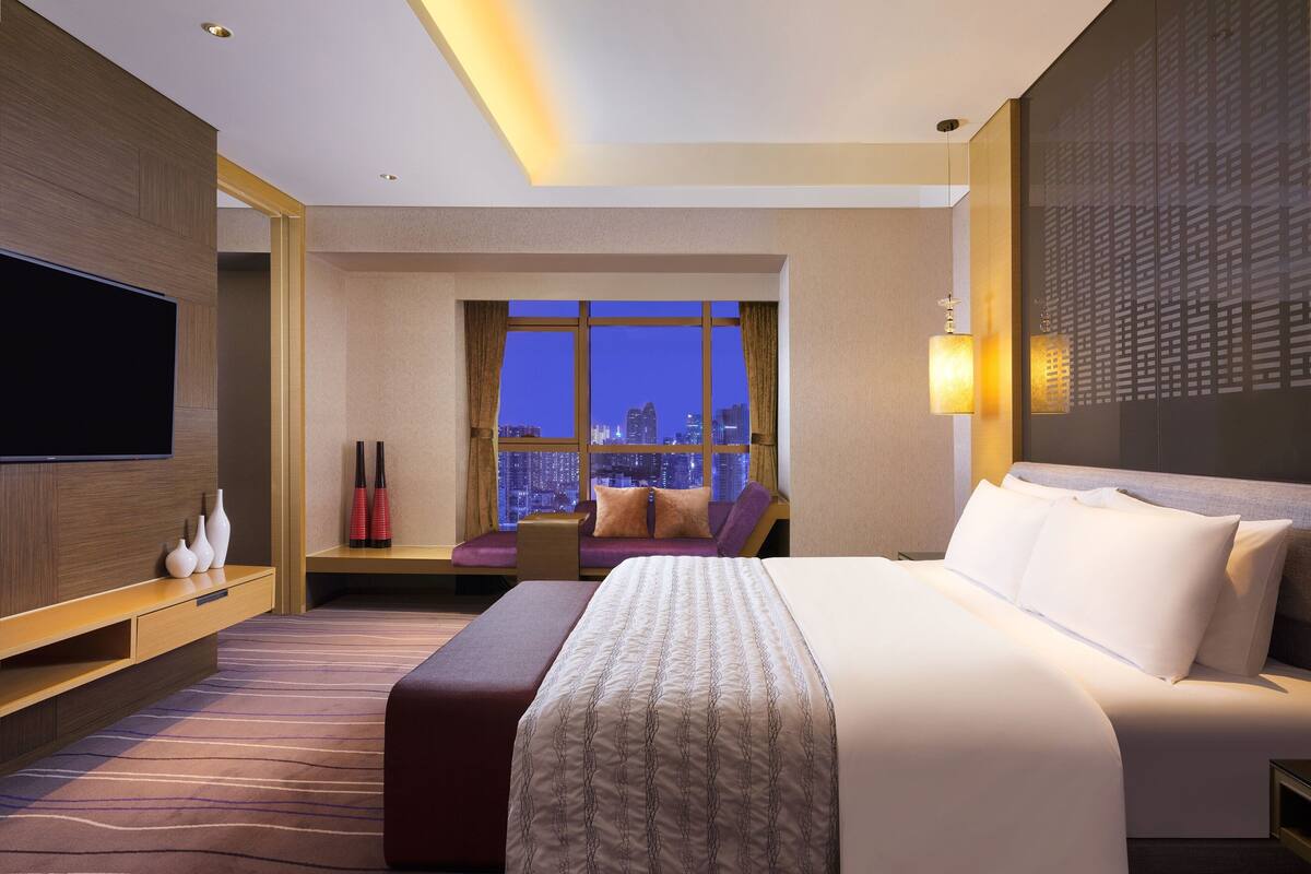 A hotel room with a large bed, a TV, a seating area with a view of the cityscape, and a city skyline visible through the window.