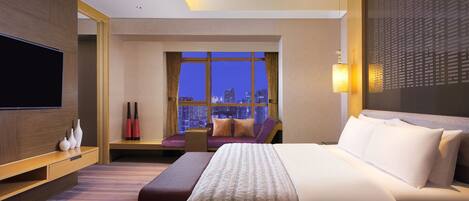 Habitación de hotel con una cama grande, televisión, zona de estar con vista a la ciudad y el horizonte urbano visible a través de la ventana.