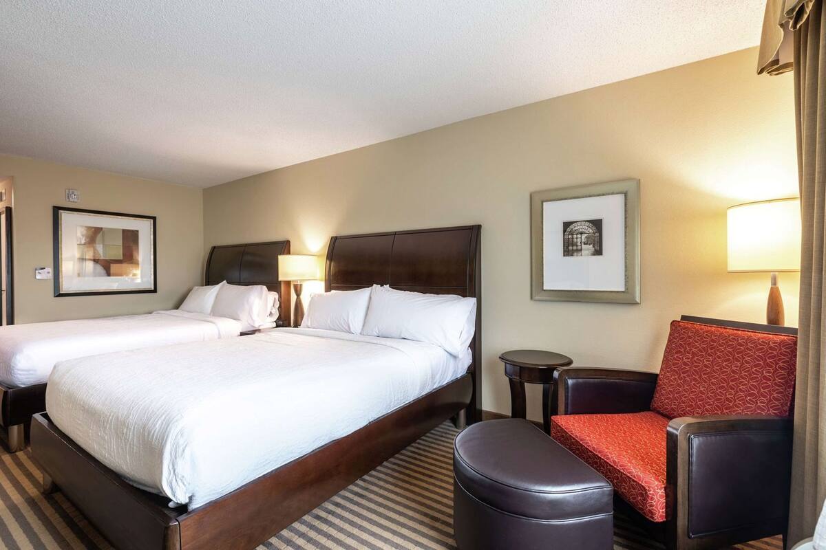A hotel room with two beds, a small table, a chair, and a lamp.