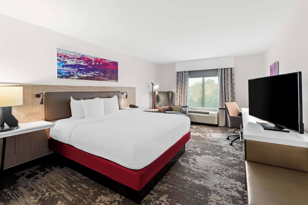Habitación de hotel con una cama grande, un televisor de pantalla plana, un escritorio con lámpara, una silla y un ventanal con cortinas.