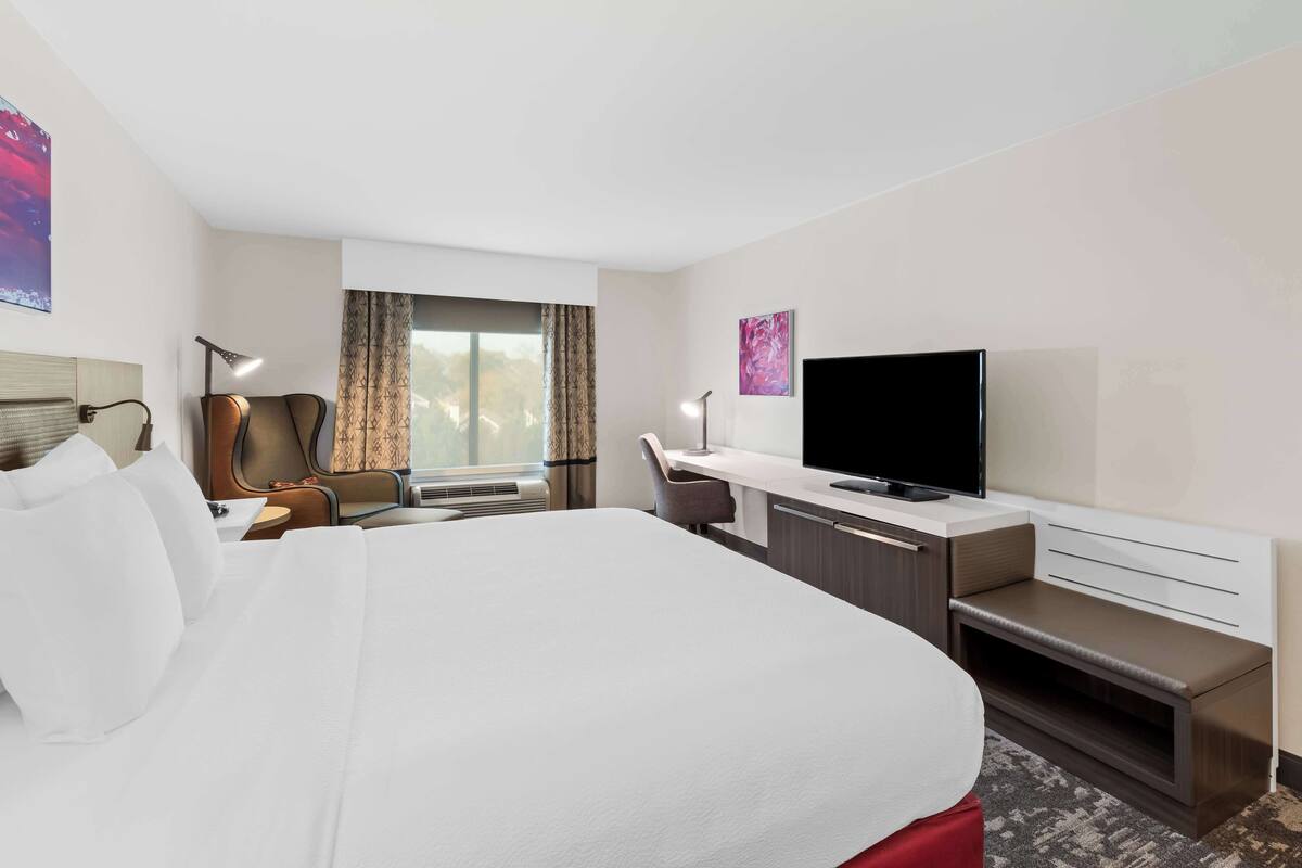 Una habitación de hotel con una cama grande, un escritorio con televisor de pantalla plana, una silla, una ventana con cortinas y un cuadro en la pared.