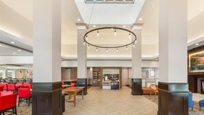 A spacious lobby with a high ceiling, a central circular light fixture, and a food service area in the background. - Hilton Garden Inn Atlanta Airport North (Atlanta)