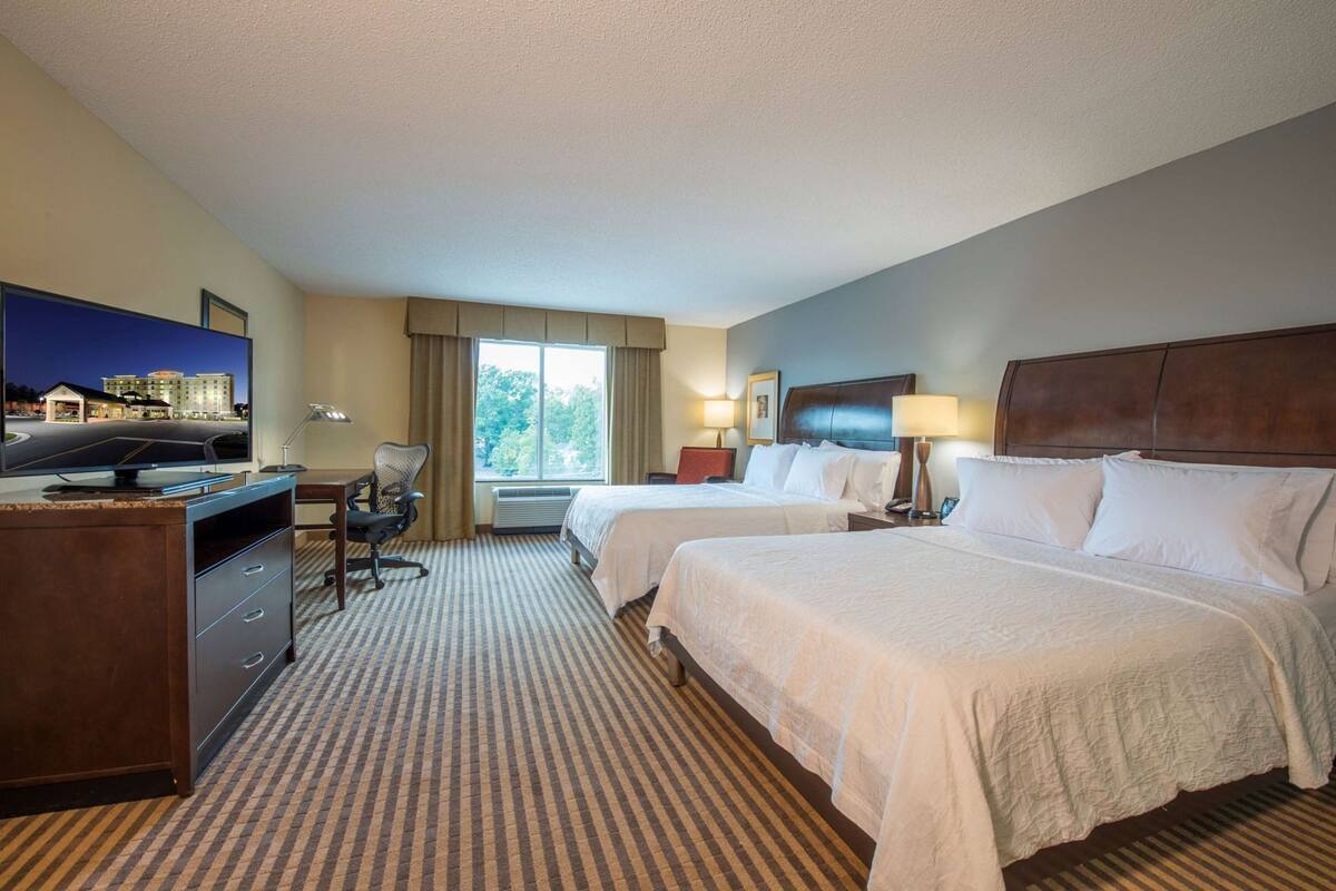 Habitación de hotel con dos camas, un escritorio con computadora, televisor y ventana con vista a edificios.