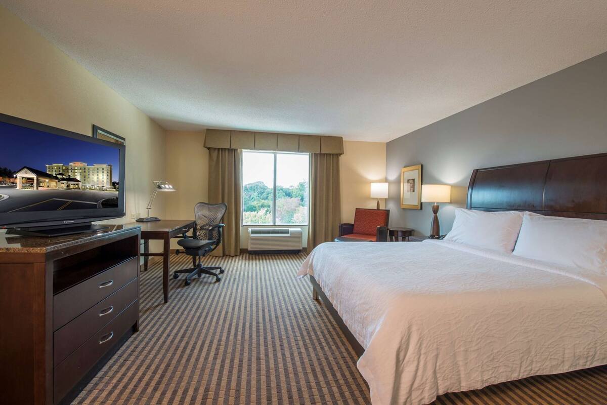 A hotel room with a bed, desk, chair, television, and a window with a view of a building.