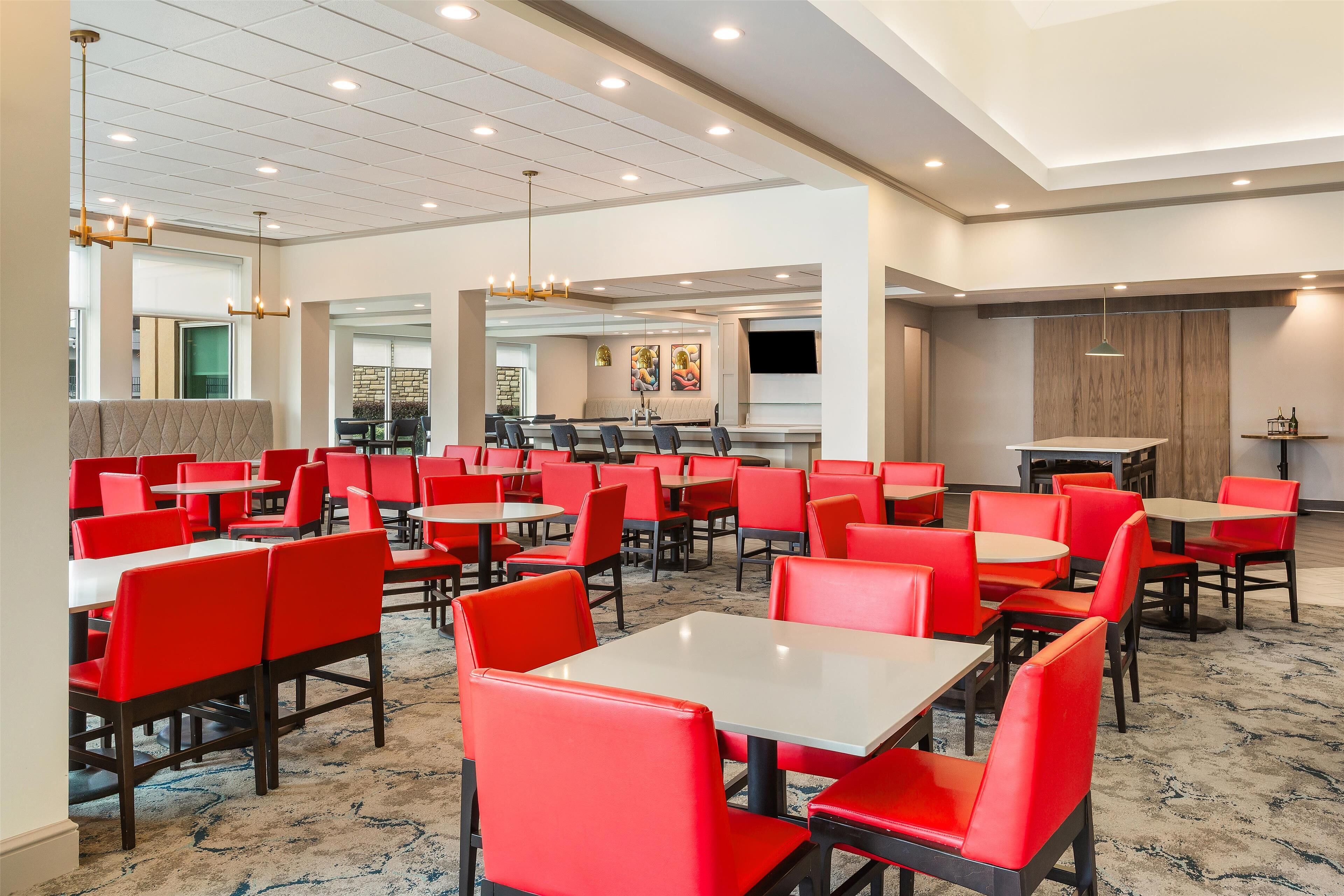 A spacious dining area with red upholstered chairs and white tables, featuring a modern ceiling design with recessed lighting and a large mirror reflecting the space.