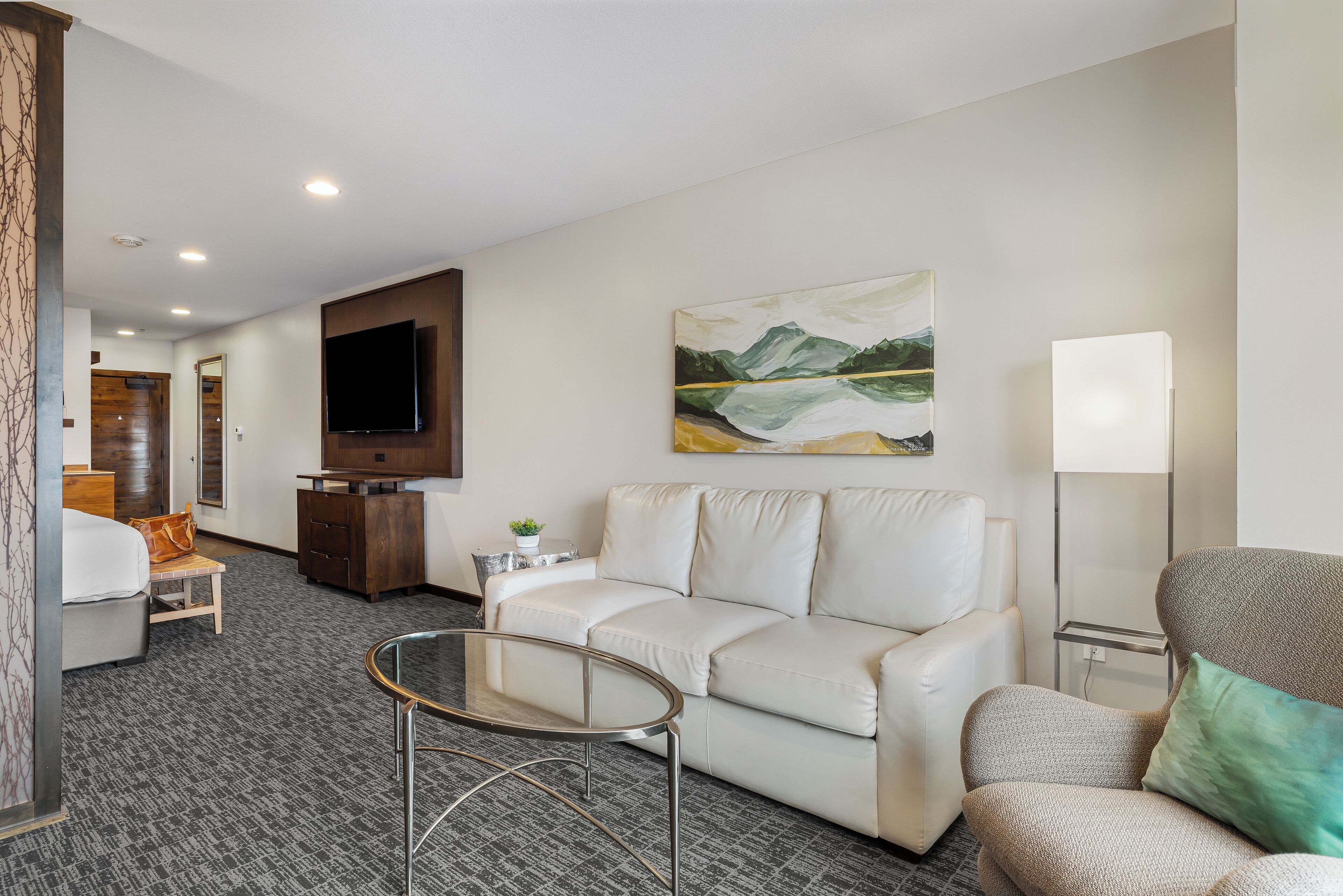A modern hotel room with a white sofa, a glass coffee table, a flat-screen TV, a painting of mountains, and a lamp.