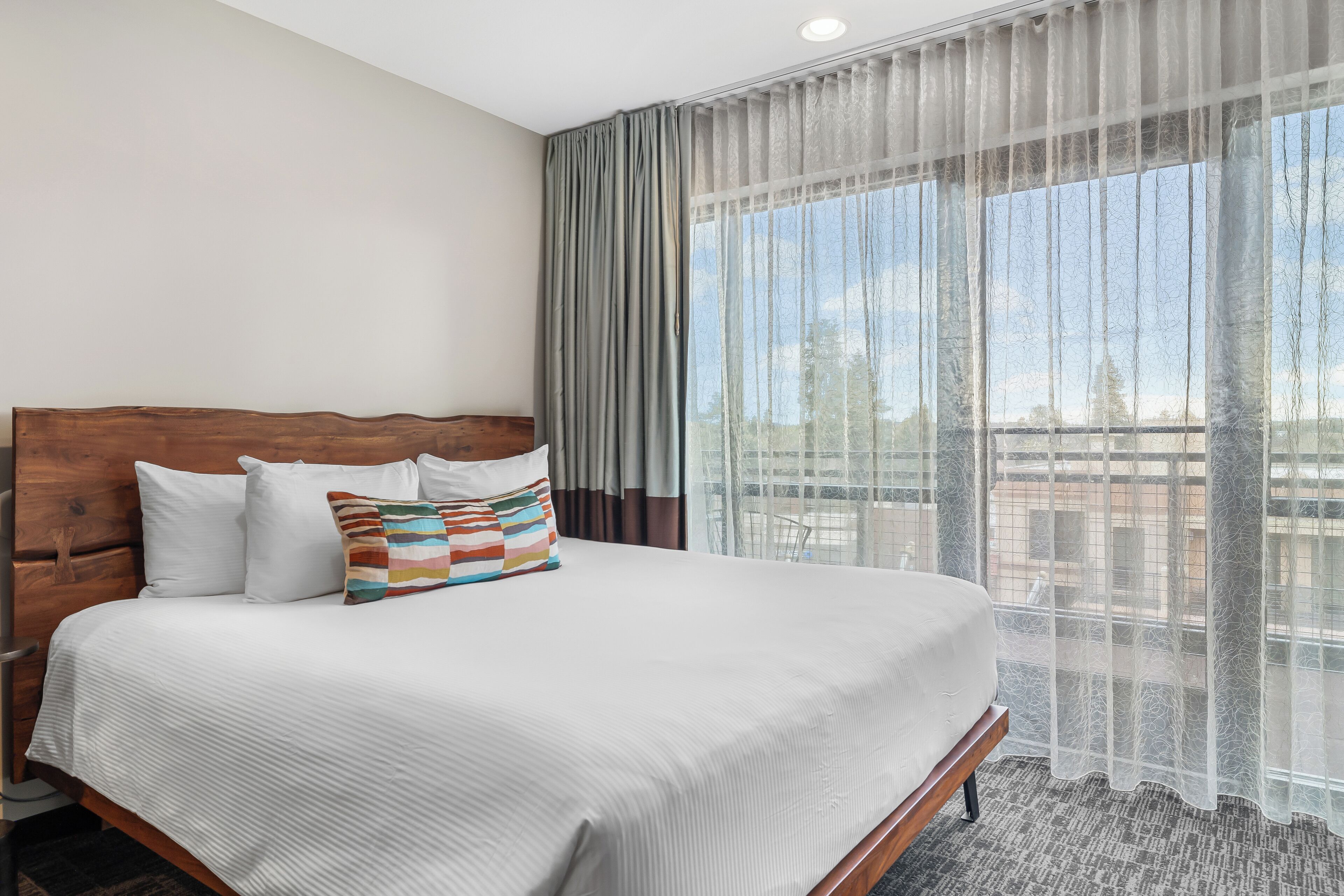 A hotel room with a large bed, a wooden headboard, white bedding, and sheer curtains.