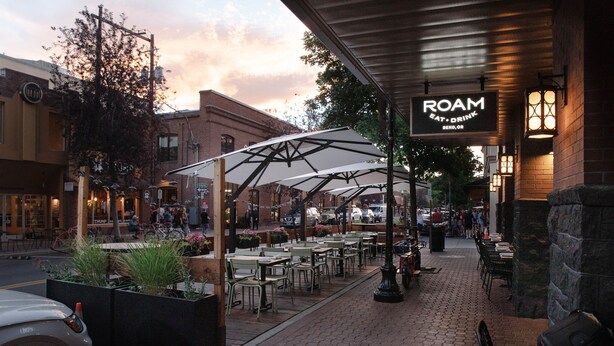 An outdoor seating area with tables and umbrellas, a sign for 'ROAM Eat + Drink', and a brick building in the background. - The Oxford Hotel Bend (Bend)