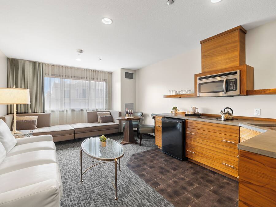 A modern living room with a sofa, a glass coffee table, and a kitchen area featuring a microwave and a refrigerator.