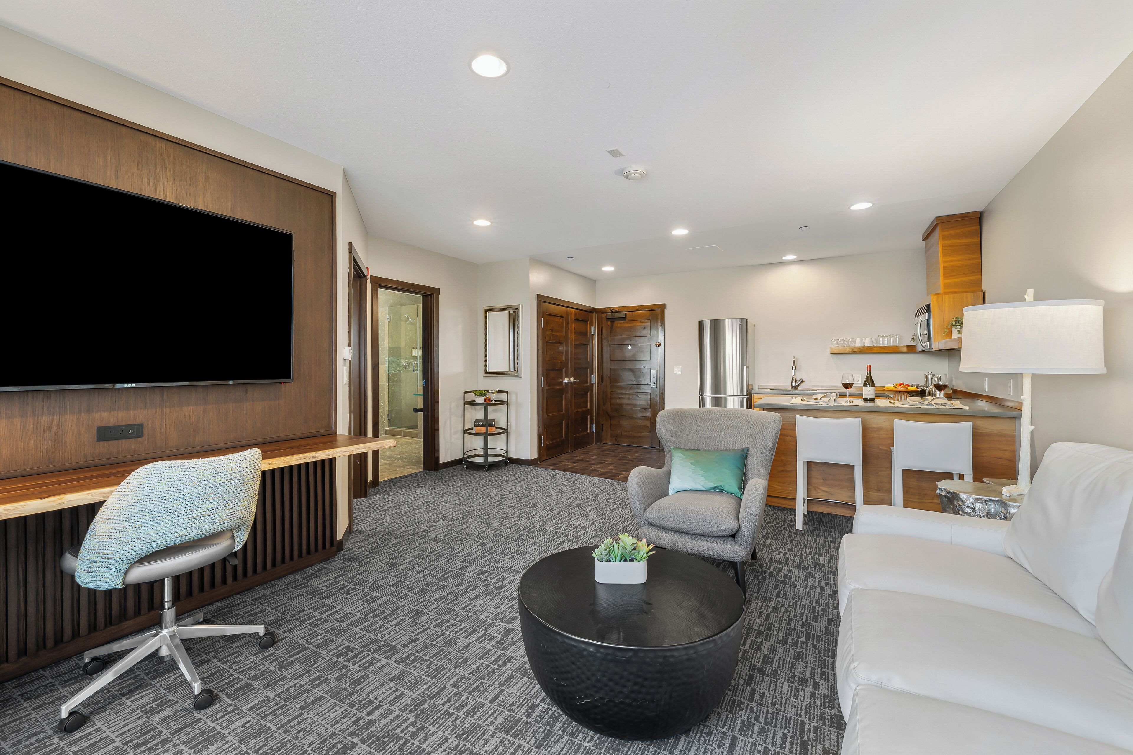 A modern hotel room with a flat-screen TV, a seating area with a sofa and armchairs, a dining area with a small table and chairs, and a kitchenette with a microwave and a lamp.
