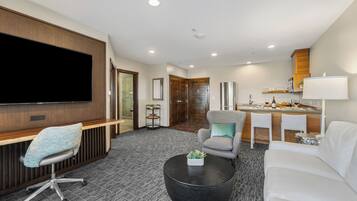 A modern hotel room with a flat-screen TV, a seating area with a sofa and armchairs, a dining area with a small table and chairs, and a kitchenette with a microwave and a lamp.